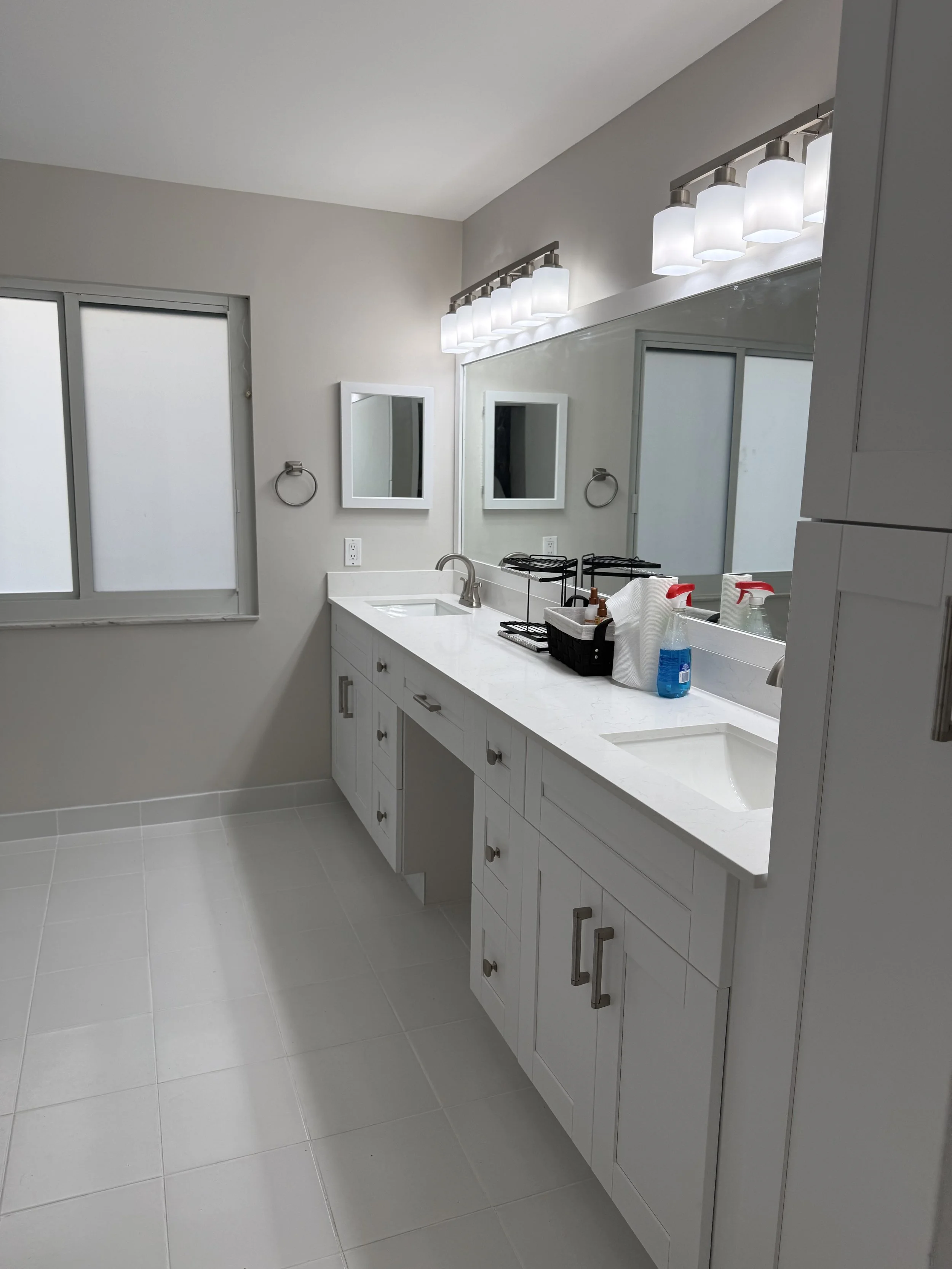 Modern white bathroom with double vanity, large mirror, frosted window, and lighting fixture with multiple bulbs