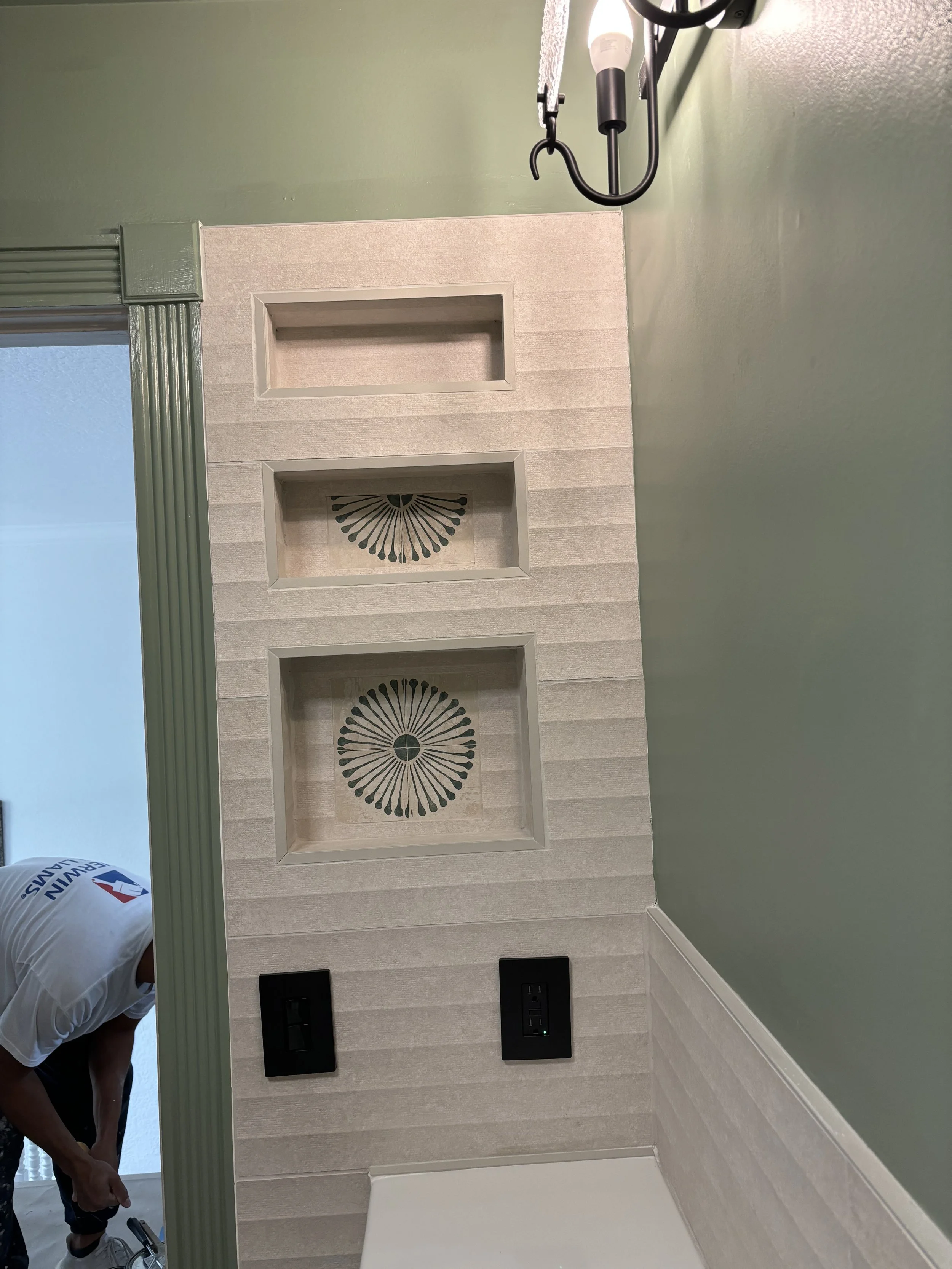 Close-up view of a bathroom backsplash with three built-in niches, decorative black fans, and two black electrical outlets, with a person's arm visible on the left side, and a green wall on the right.