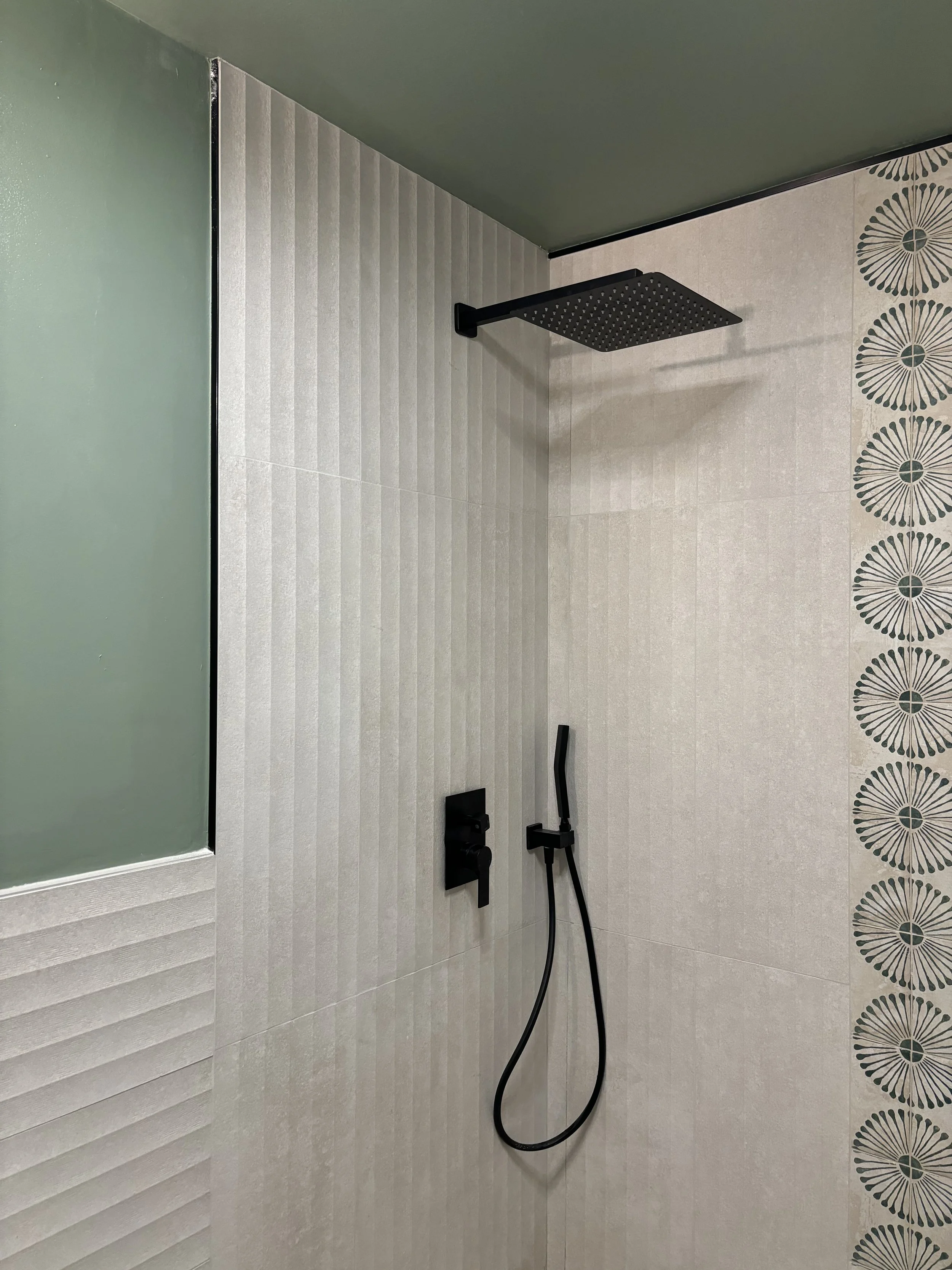 Modern shower area with a black rain shower head, handheld shower wand, and black fixtures against a tiled wall with a decorative border pattern.