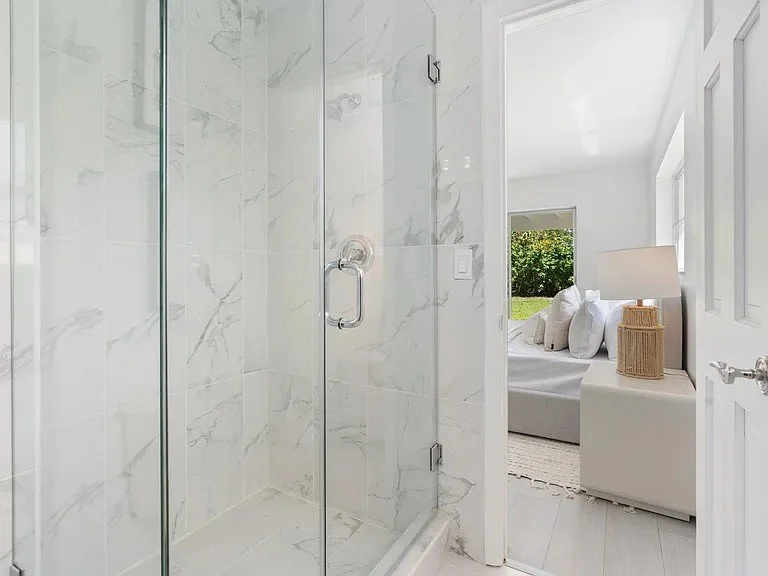 Modern bathroom with a glass shower and a view into a bright bedroom with a large window, white bedding, and a nightstand with a lamp and decorative basket.