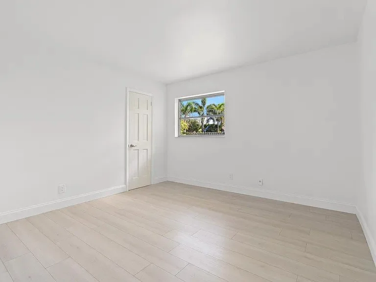 Empty white room with a window showing palm trees outside and a closed door.