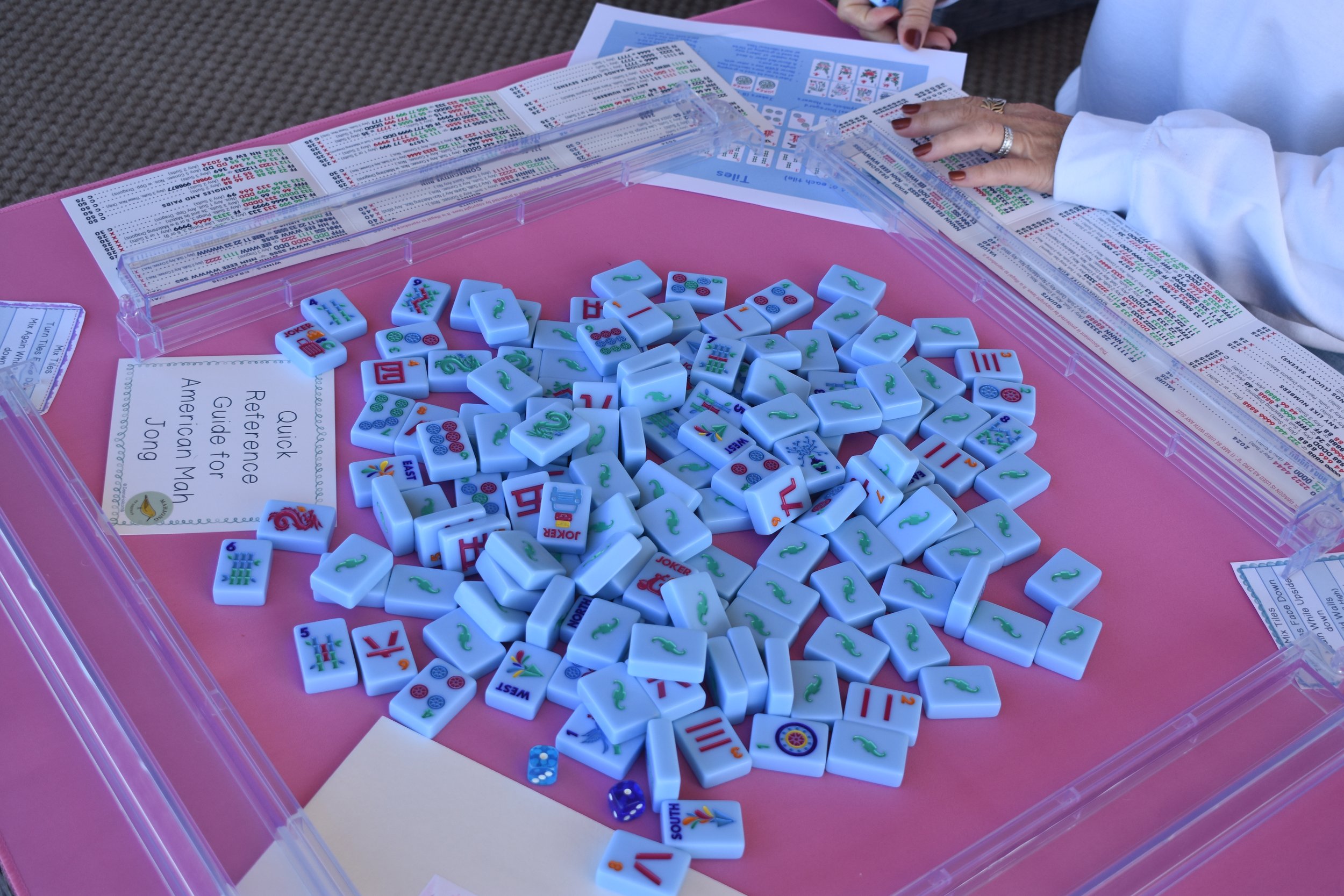 A mahjong game setup on a pink table with scattered mahjong tiles, a plastic rack, a pair of blue dice, and printed guides. A person with painted nails and rings is reaching towards the tiles.