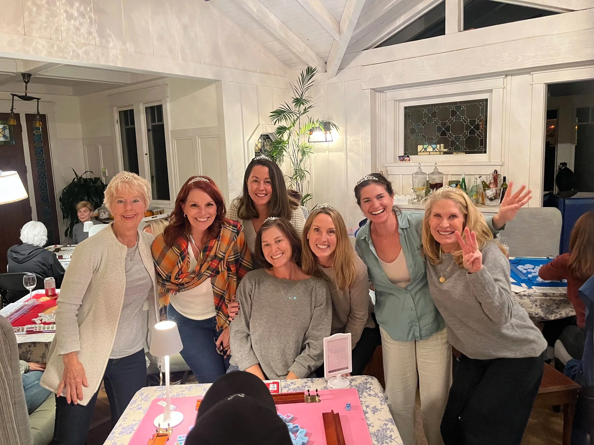 Group of women smiling and posing at a social gathering, some women wearing tiaras, in a well-lit indoor space with a white wooden ceiling, plants, and a table with game pieces.