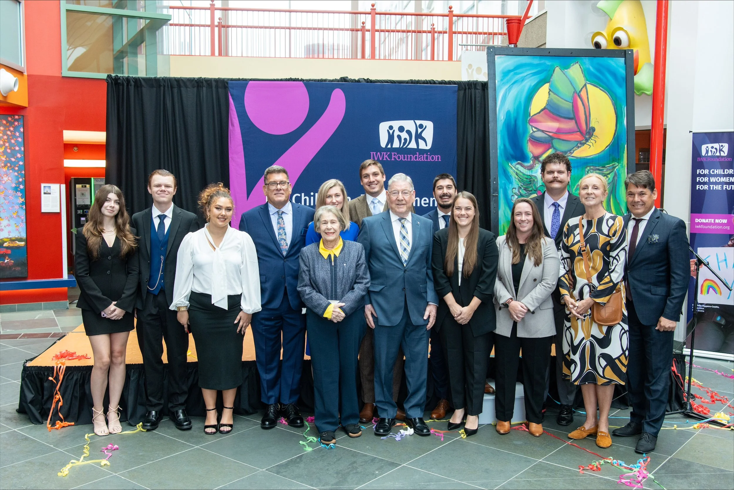 Group of people posing for a photo in front of a black curtain with the IW K Foundation logo during an event. The group includes men and women dressed in formal attire, standing on a decorated stage with colorful streamers on the floor.