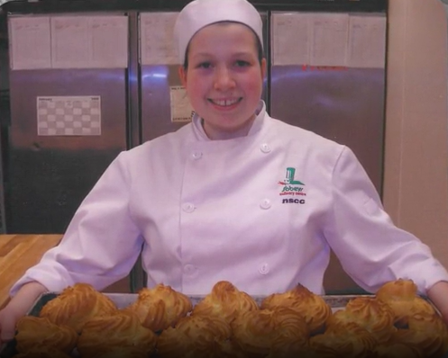 A young girl dressed as a chef, smiling, displaying a tray of baked pastries in a kitchen.