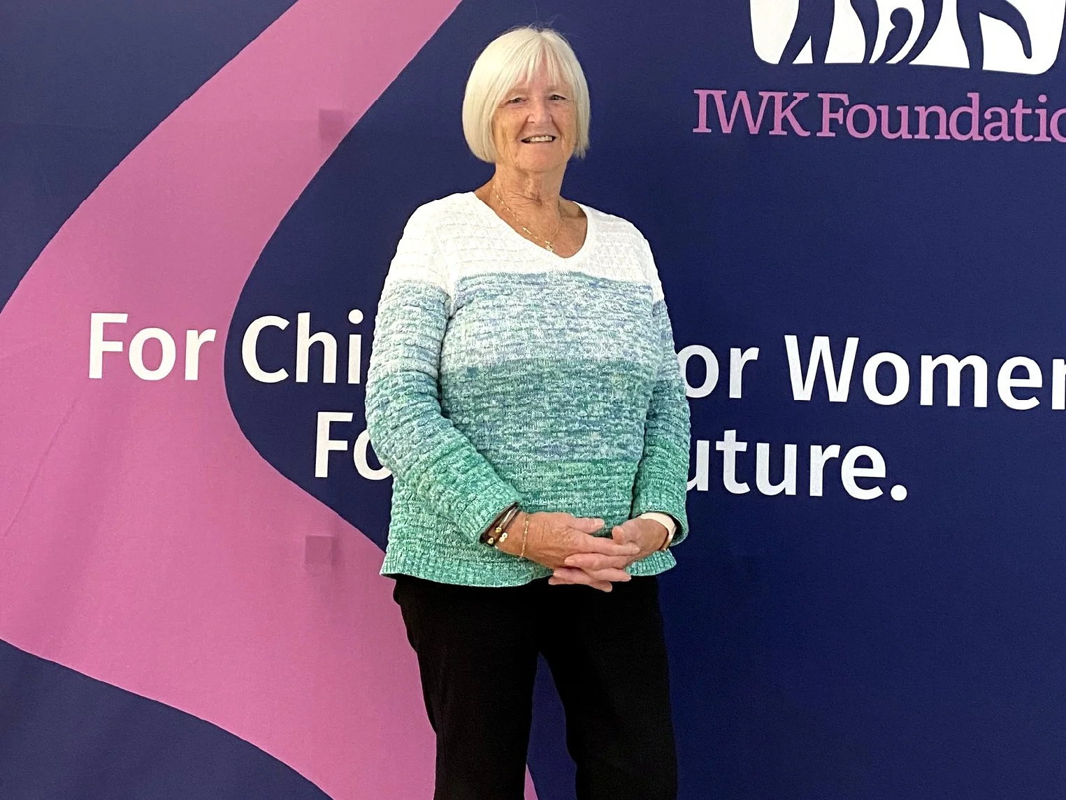 An elderly woman with short blonde hair smiling, wearing a multicolored sweater and black pants, stands in front of a pink and blue backdrop with the logos and text related to the Susan G. Komen Foundation for breast cancer awareness.