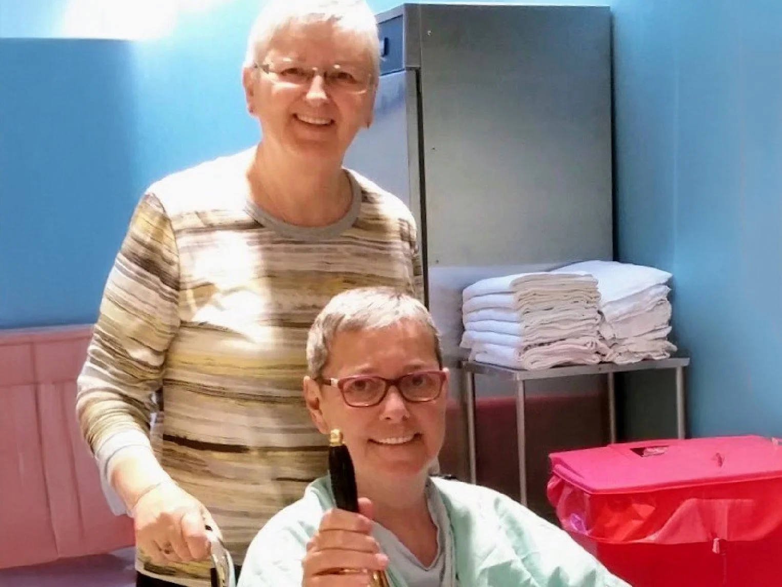 Two women in a room with blue walls, one sitting and one standing behind her. The seated woman has short gray hair, wears glasses and a medical gown, and holds a bottle. The standing woman has short blond hair, glasses, and is wearing a striped long sleeve shirt. There are folded towels on a table and a large red container nearby.