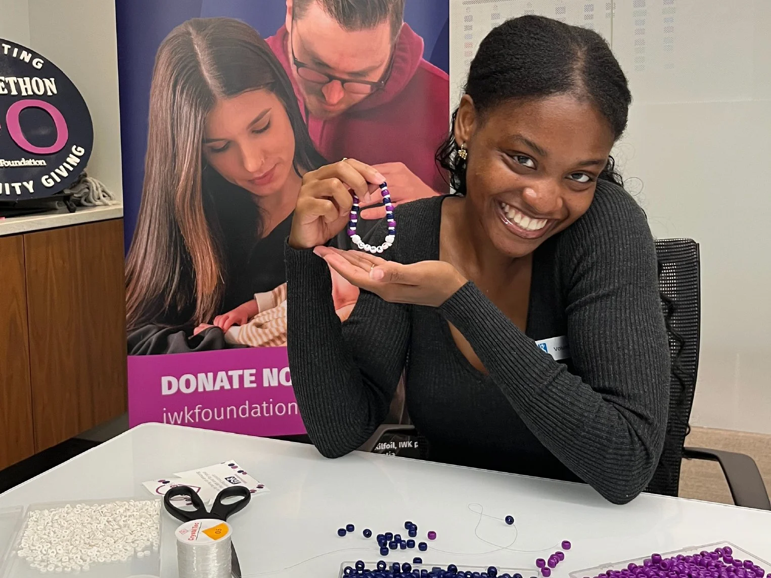 A smiling woman with dark hair and a black sweater, holding a beaded bracelet she made, sitting at a table with beads, scissors, and thread. Behind her is a poster promoting a foundation and a photo of two people looking at beads.