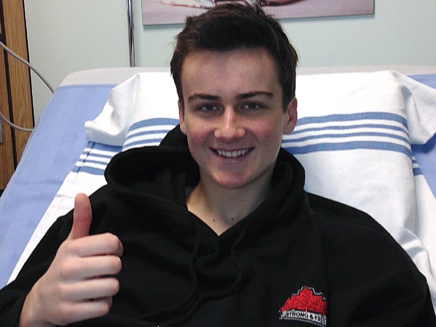 Young man named Curtis Kennedy smiling and giving a thumbs up while lying on a hospital bed at the IWK