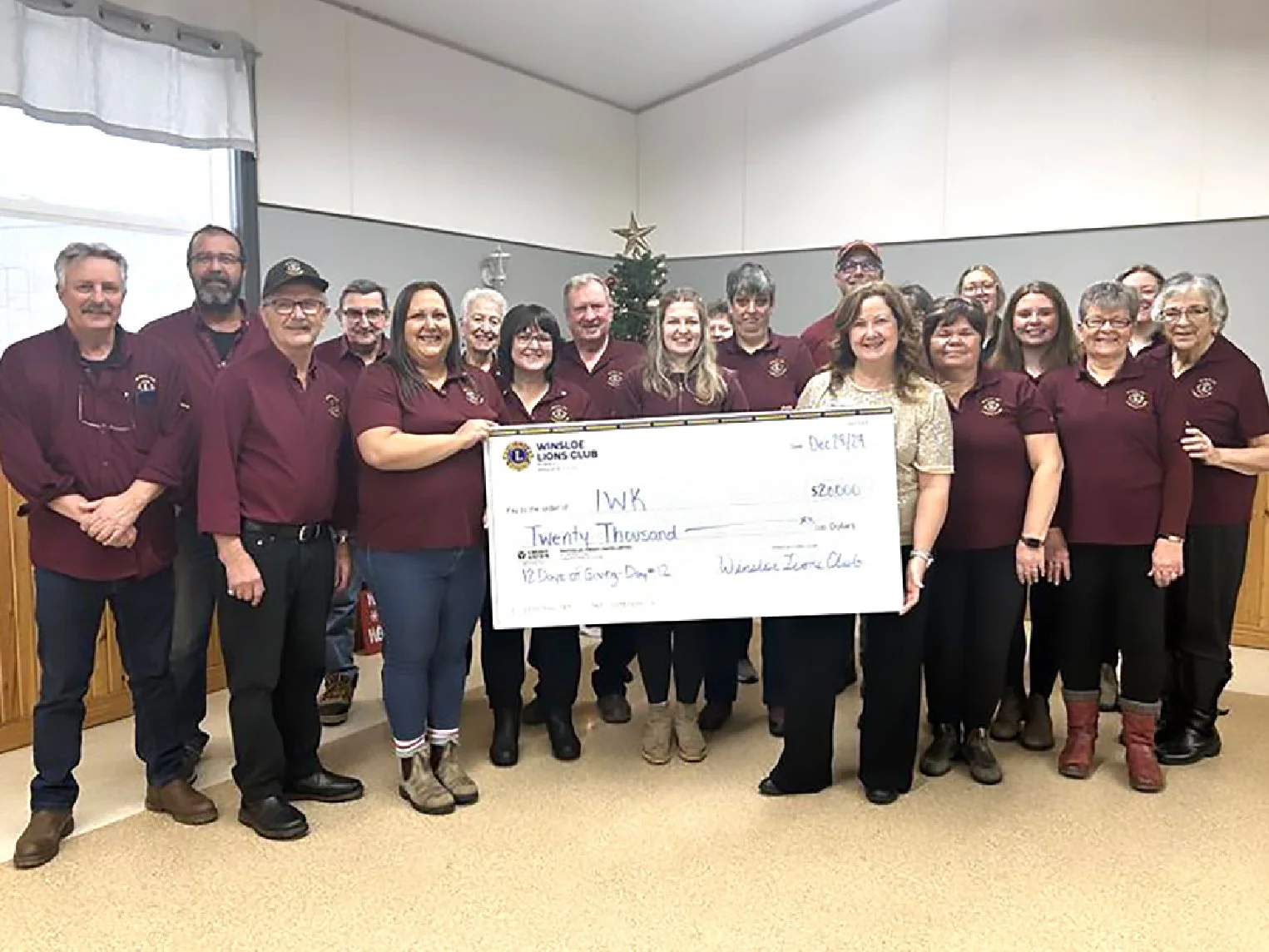 Group of people wearing maroon shirts holding a large check for twenty thousand dollars in a room decorated for Christmas, with a small Christmas tree visible in the background.