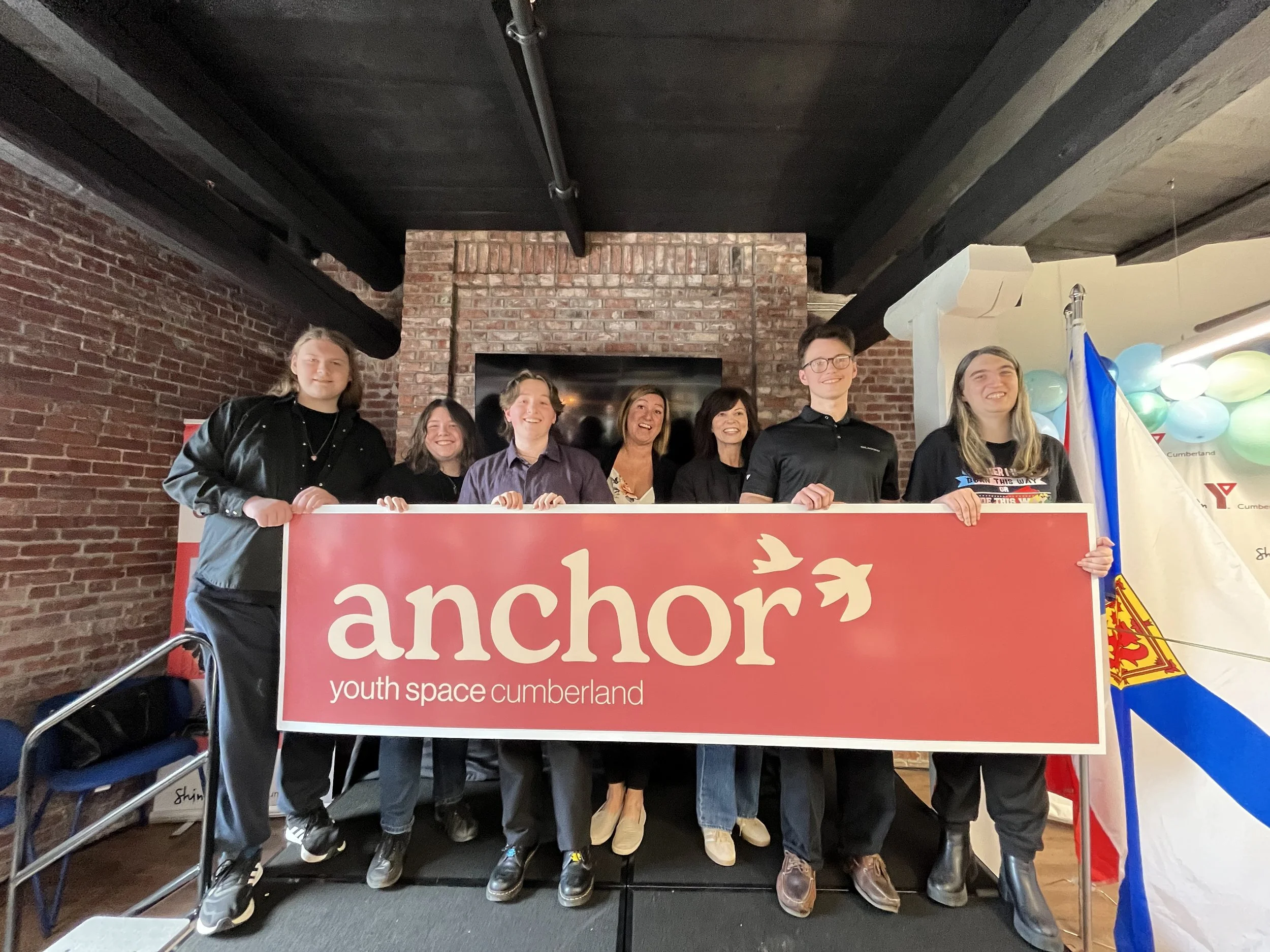 Group of young people holding a red banner that reads 'anchor youth space cumberland' at a YMCA event, with balloons and flags in the background.