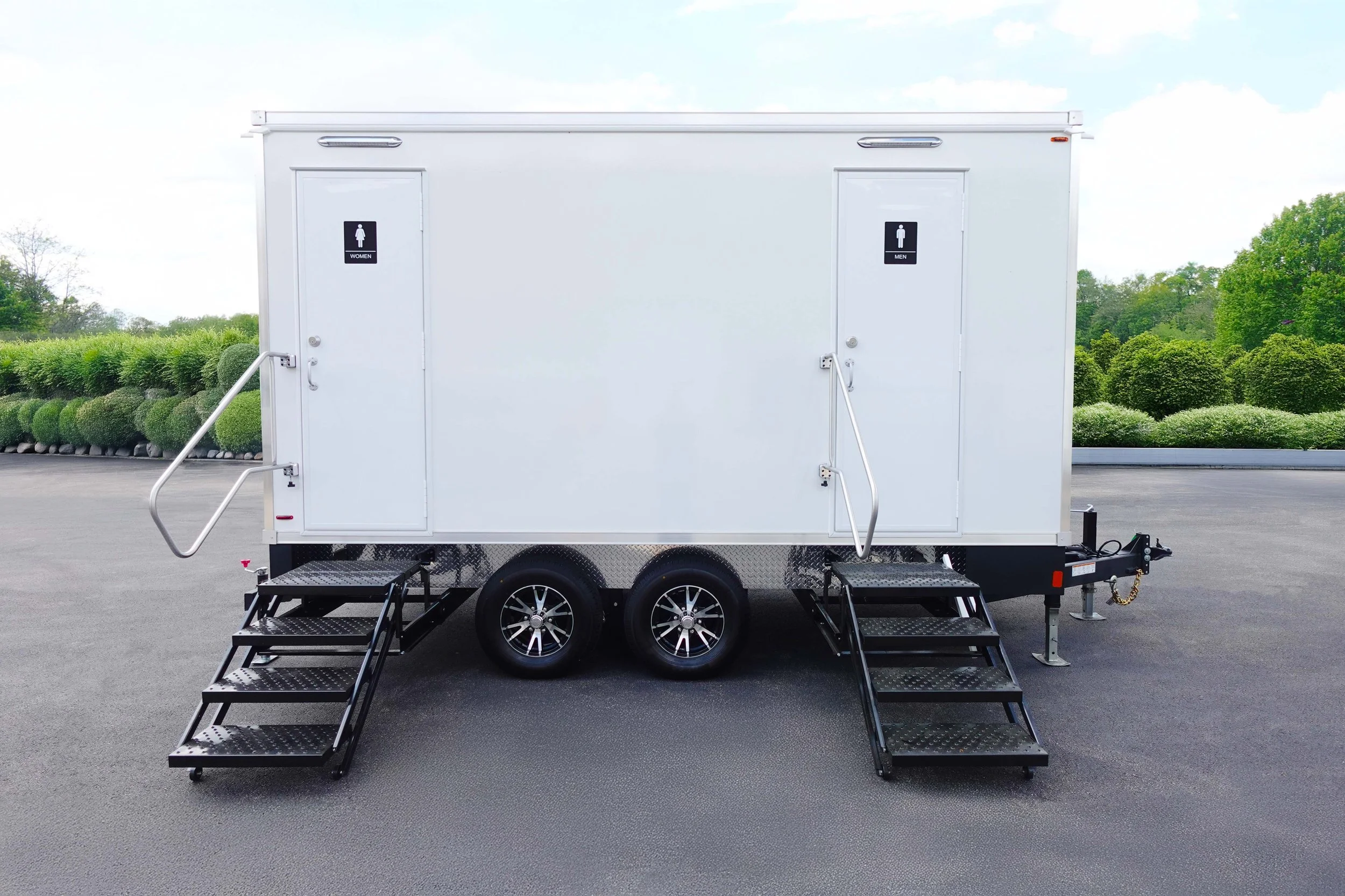 A white mobile restroom trailer with two doors, one labeled for women and one for men, and metal stairs leading up to each entrance, parked on an asphalt lot with green bushes and trees in the background.