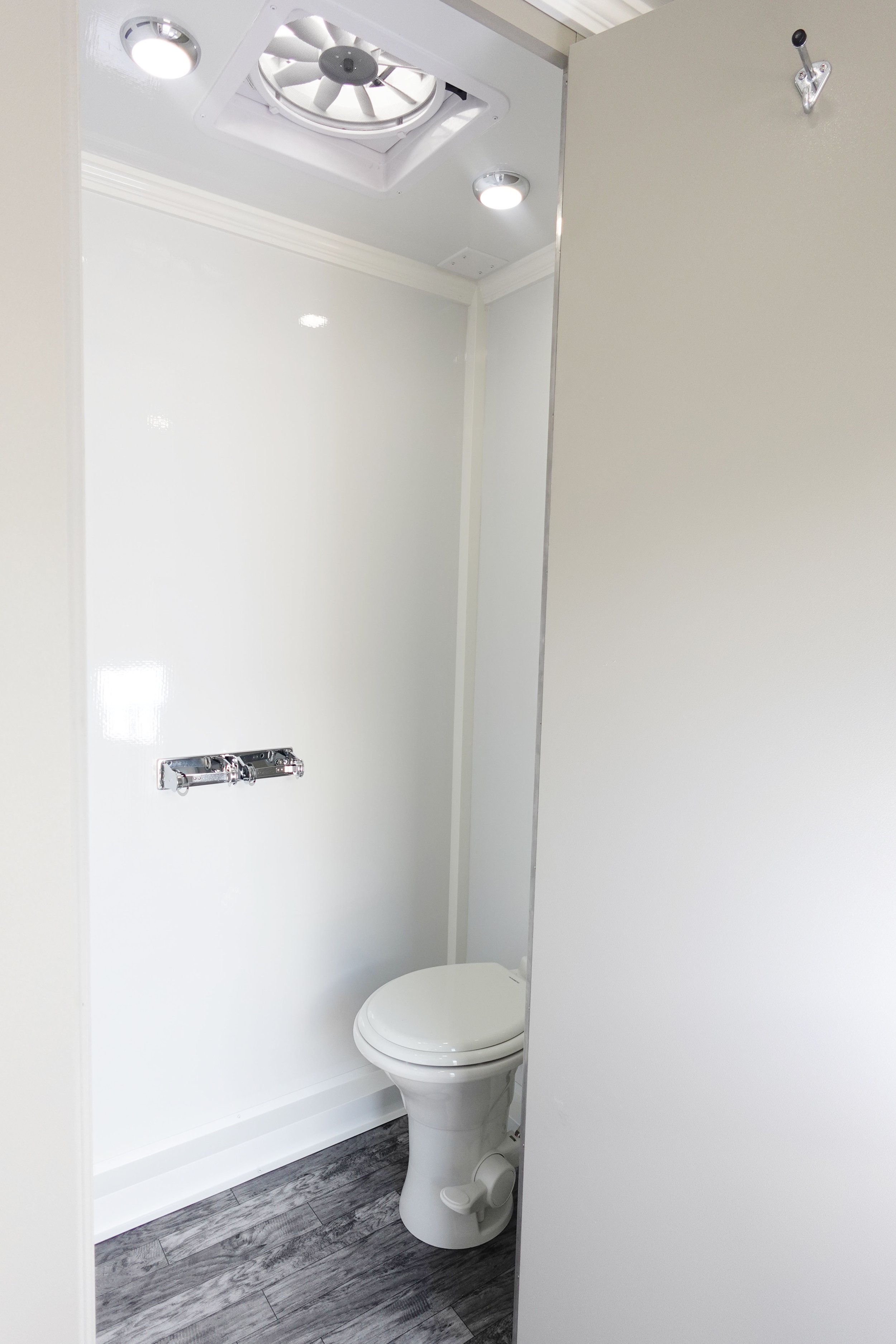 Small bathroom with white walls, a white toilet, a wall-mounted paper holder, a ceiling fan, and a floor with dark wood planks.