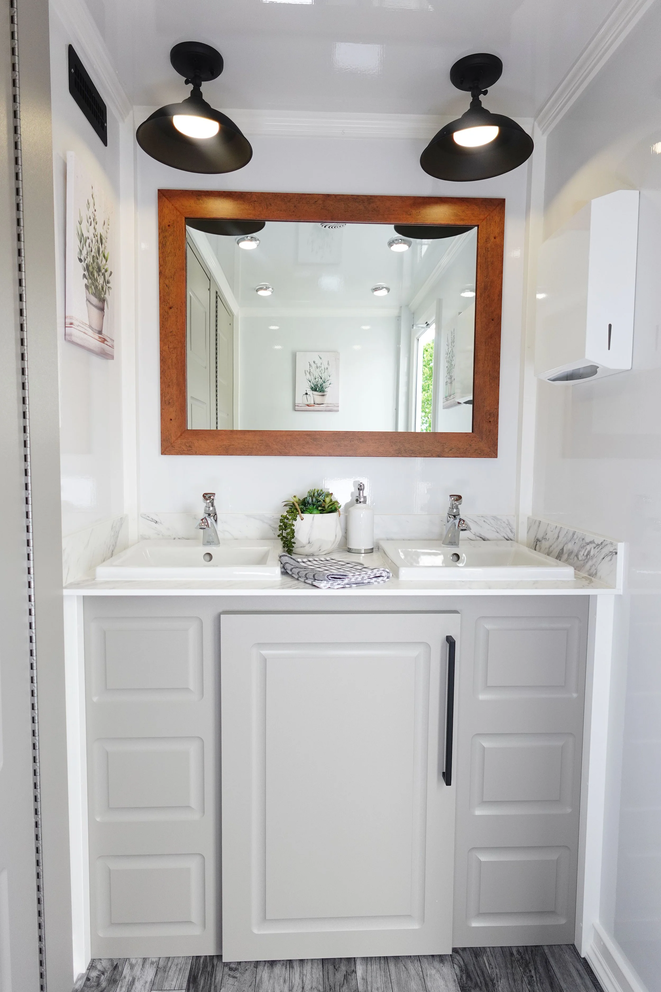 Bathroom vanity with dual sinks, a large mirror, two black ceiling lights, and small plants on the countertop.