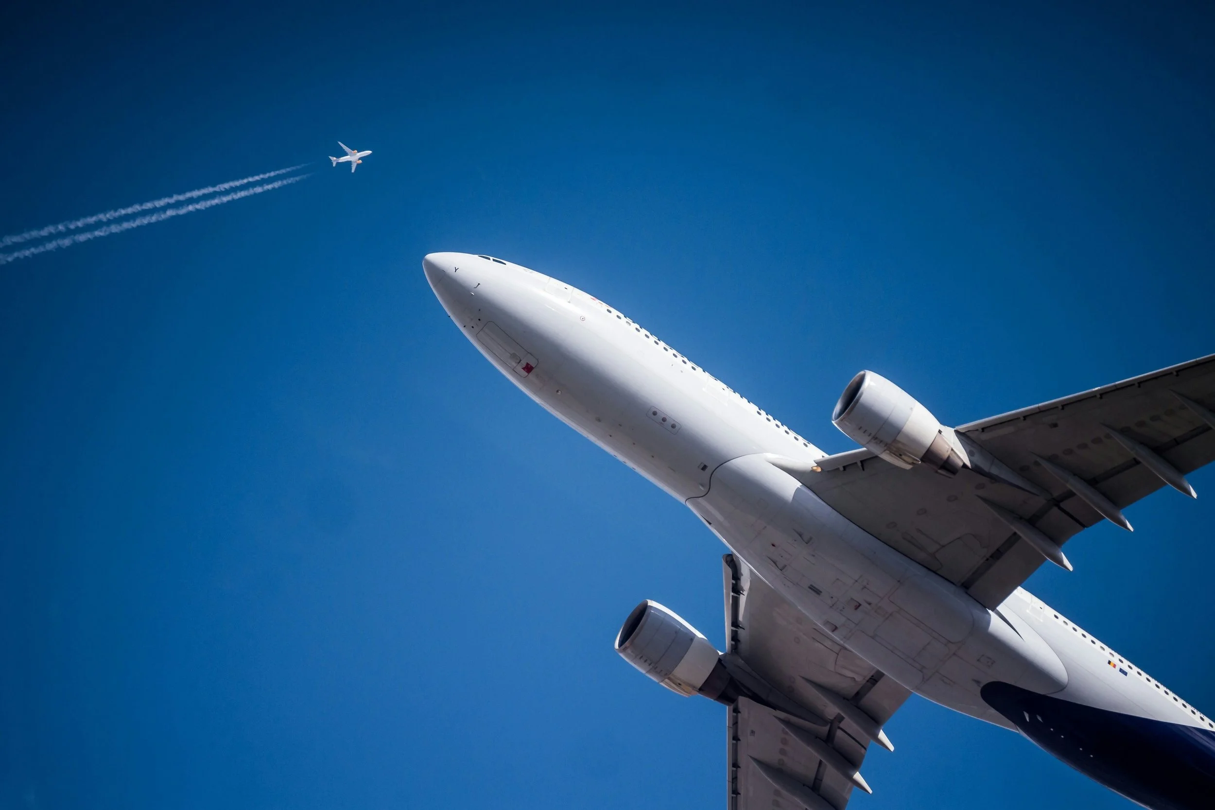 A large white commercial airplane flying in the clear blue sky with a smaller airplane passing by in the background, leaving contrails.