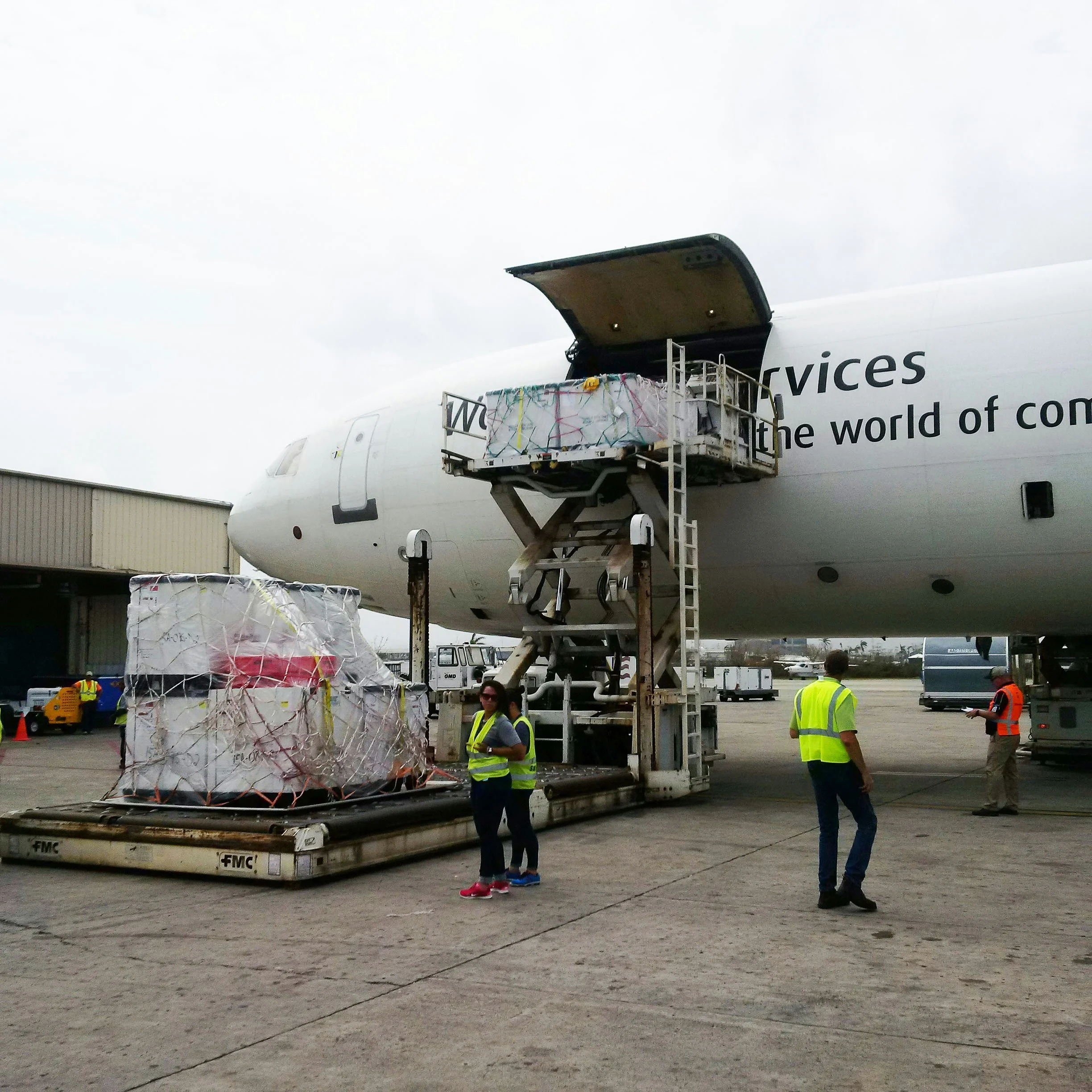 Cargo being loaded onto an airplane at an airport, with ground crew and cargo pallets visible.