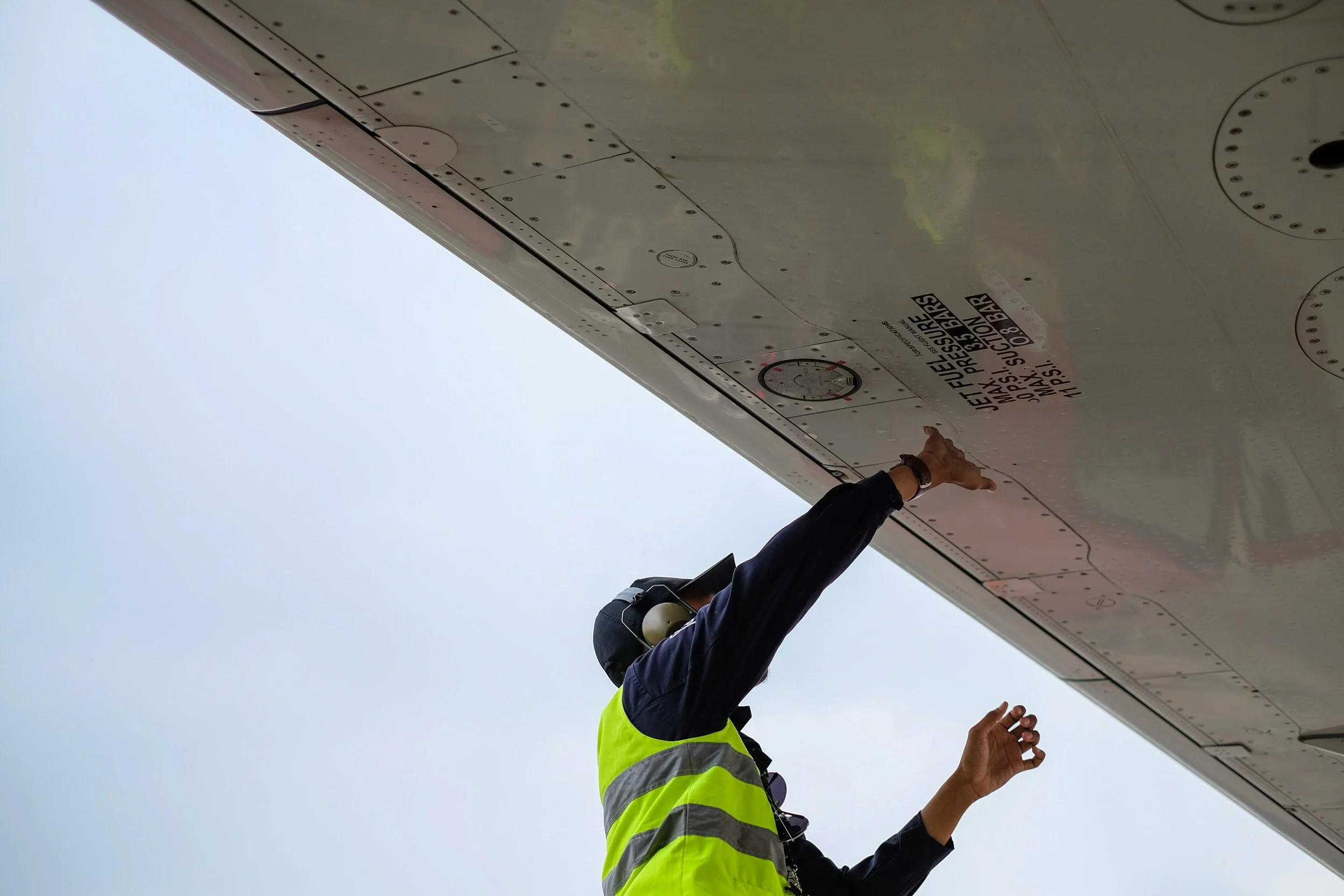 A person in a high visibility vest and a black cap inspecting aircraft's underside, pointing towards a panel on the aircraft's wing.