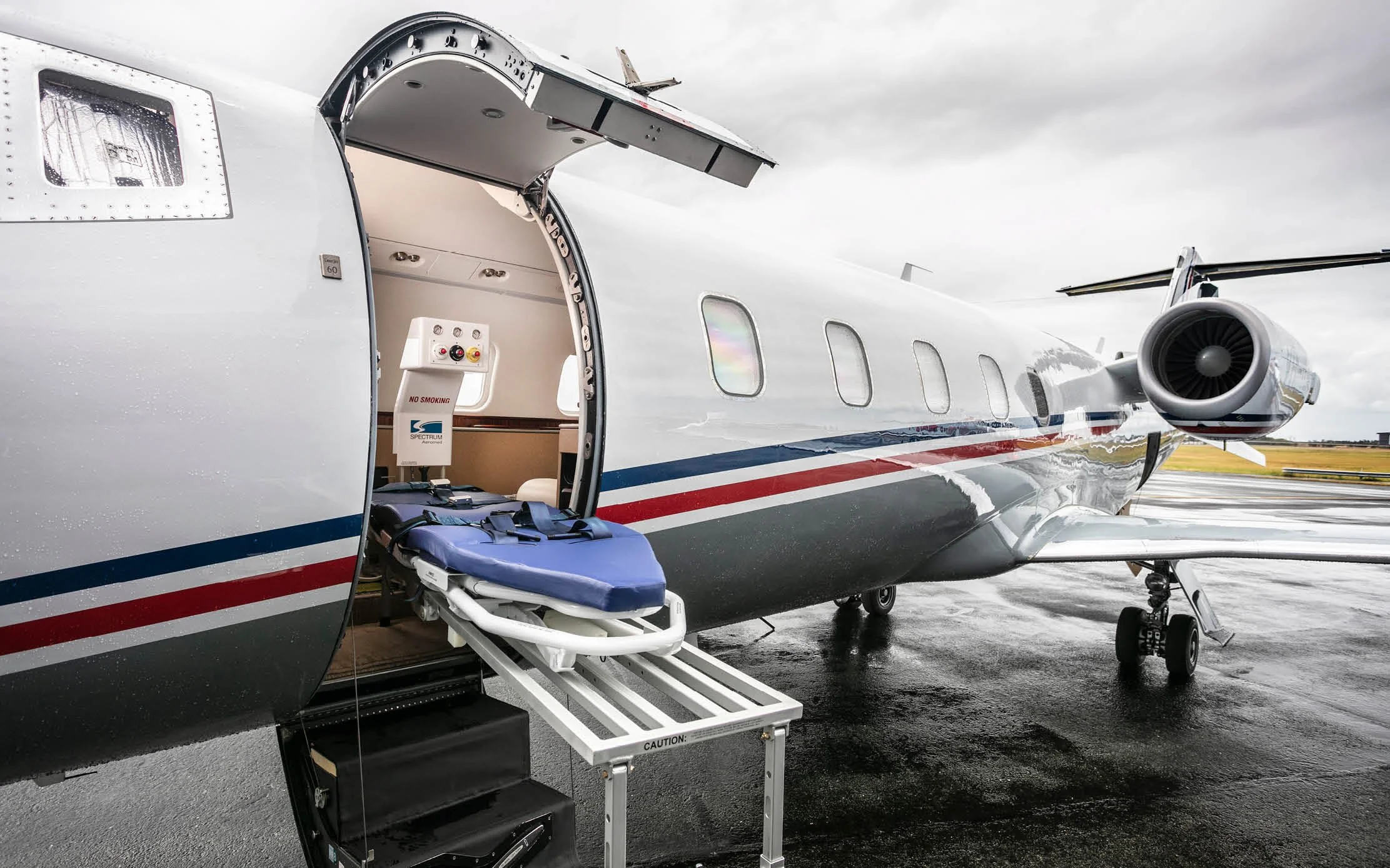 Small private jet on tarmac with open door, medical stretcher inside, and a cloudy sky overhead.