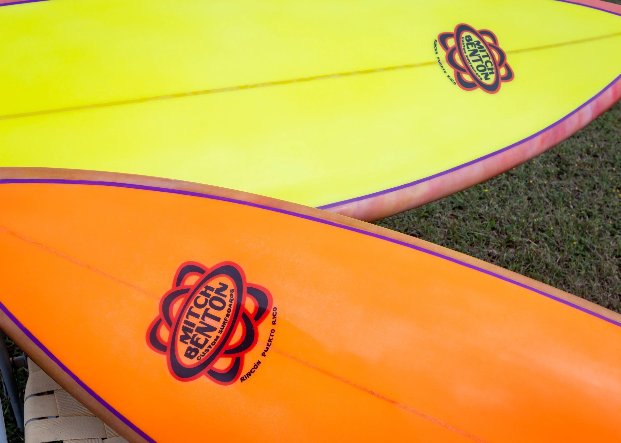 Close-up of two surfboards, one yellow and one orange, with a 'Mitch Bennetton' logo and 'Rincon Puerto Rico' text on each, placed on grass.