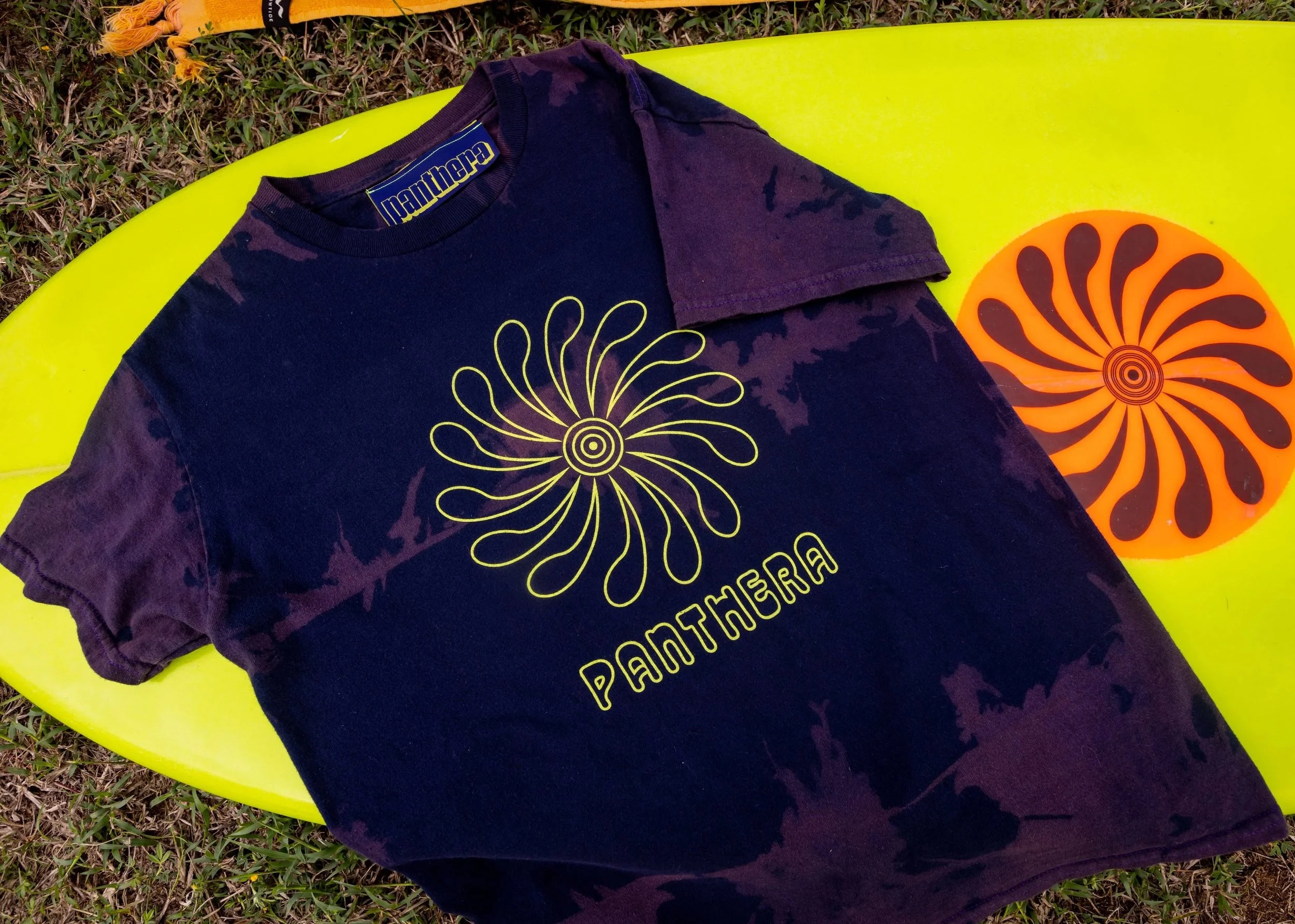 A black t-shirt with yellow graphic and text, laying on a yellow surfboard with orange circular design. The t-shirt has a stylized yellow spiral flower design and the word 'PANTHERA' printed below the flower. The surfboard is on grass, and there's a yellow leash attached at the top of the surfboard.