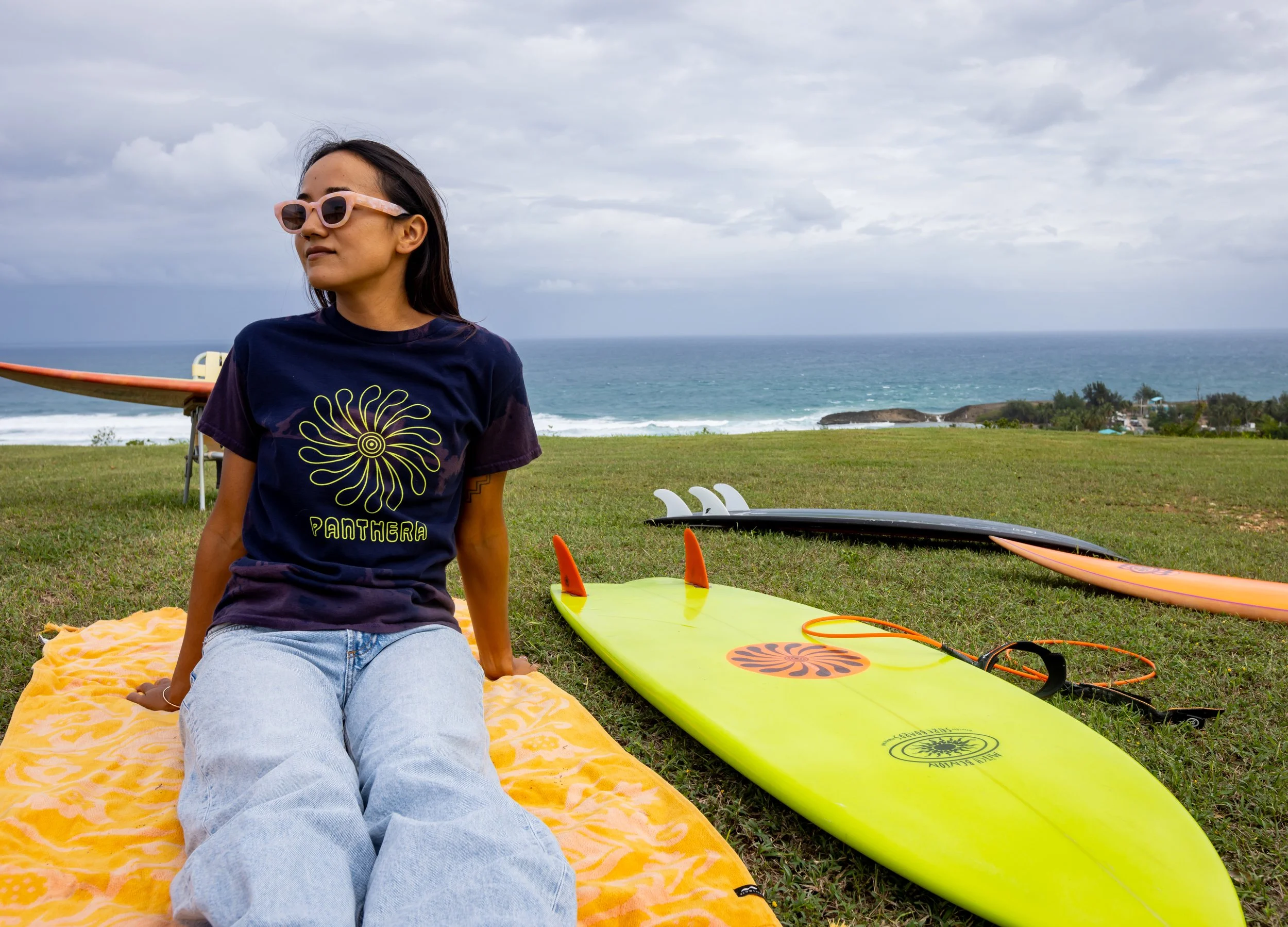 A woman sitting on a yellow towel on grass near the beach, wearing sunglasses and a t-shirt with the word 'PANTHERA' and a spiral sunflower design, with surfboards lying on the grass nearby and the ocean in the background.