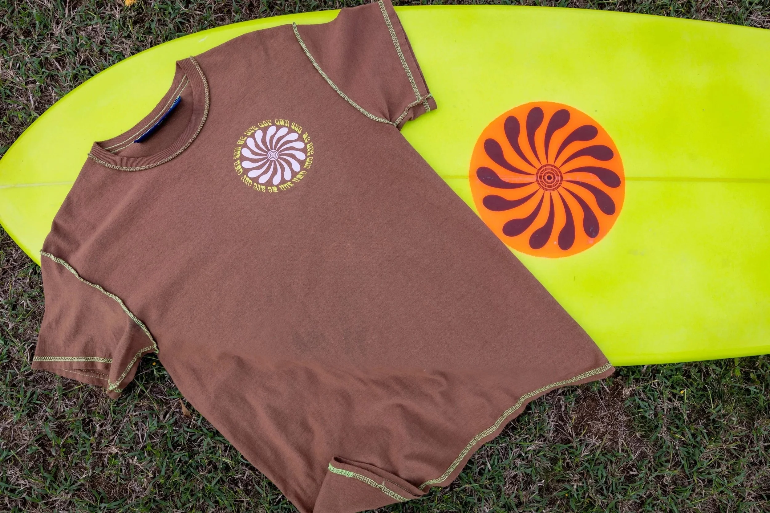 A brown T-shirt with a spiral and sunburst design, and a surfboard with a matching orange and black sunburst sticker, lying on grass.