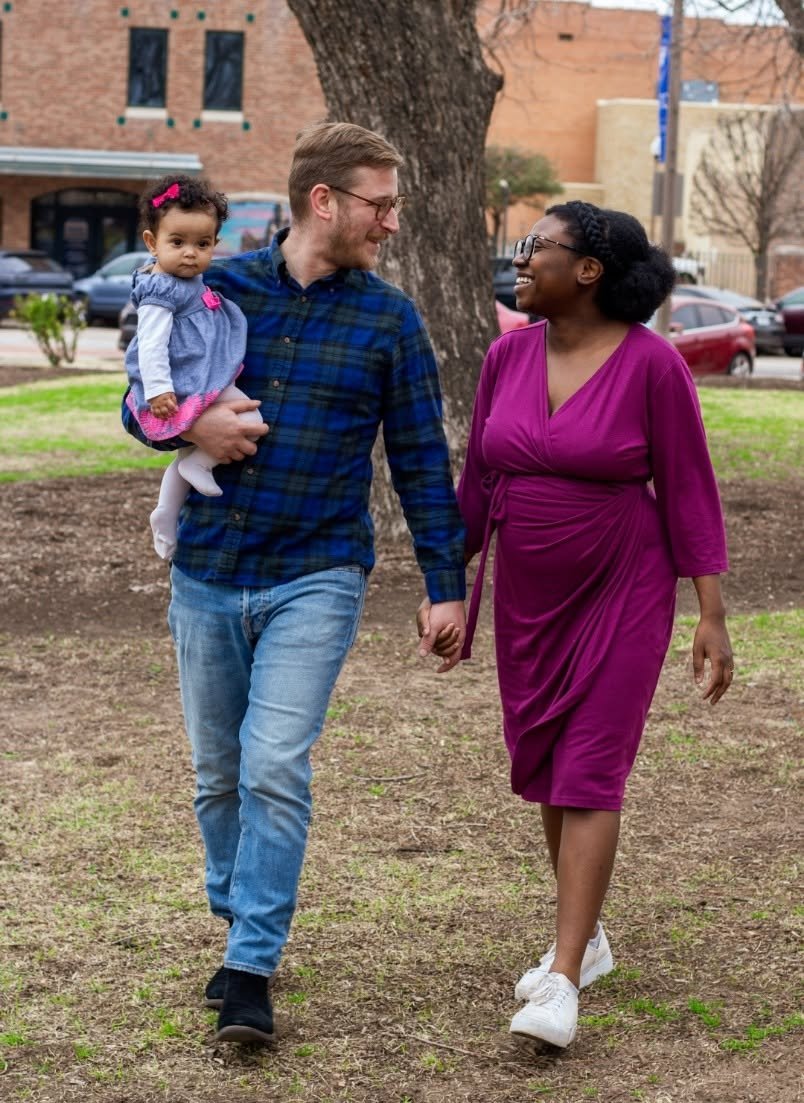 A family of three walking outdoors, holding hands, with a man carrying a young girl on his shoulder, a woman in a purple dress, and a large tree in the background.