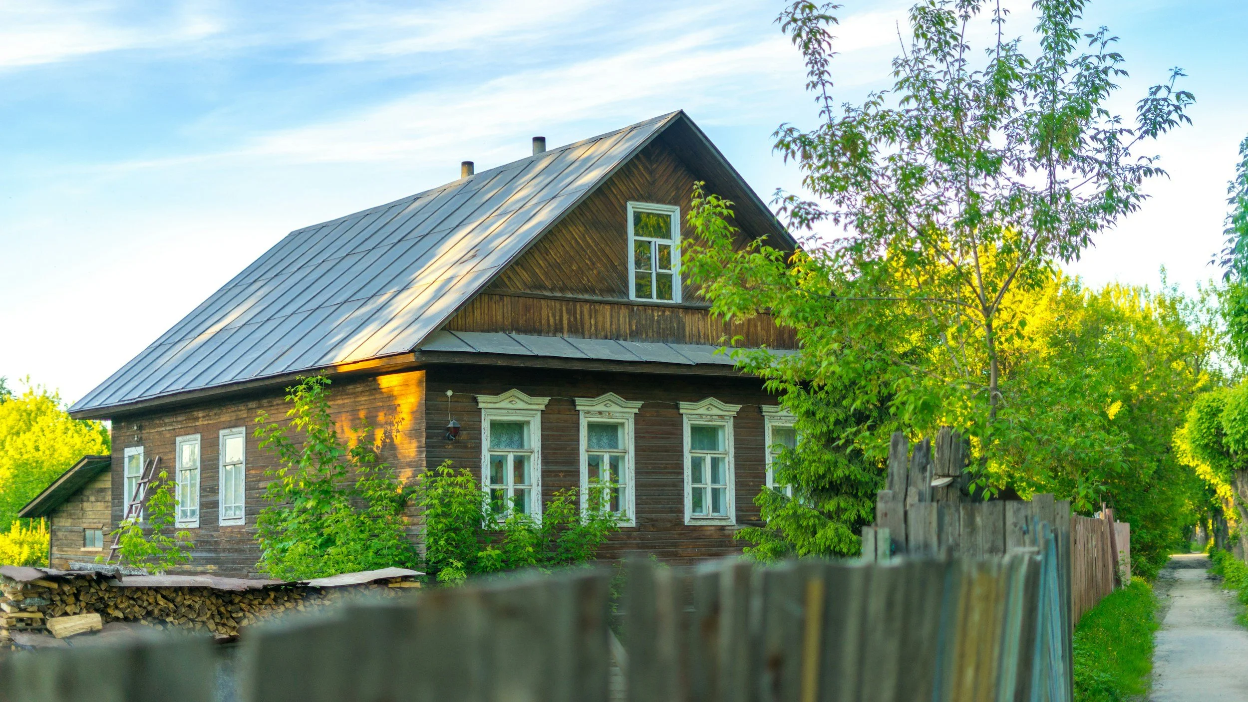 A wooden house with white window frames surrounded by green trees and plants, with a weathered wooden fence in the foreground, under a blue sky with light clouds.