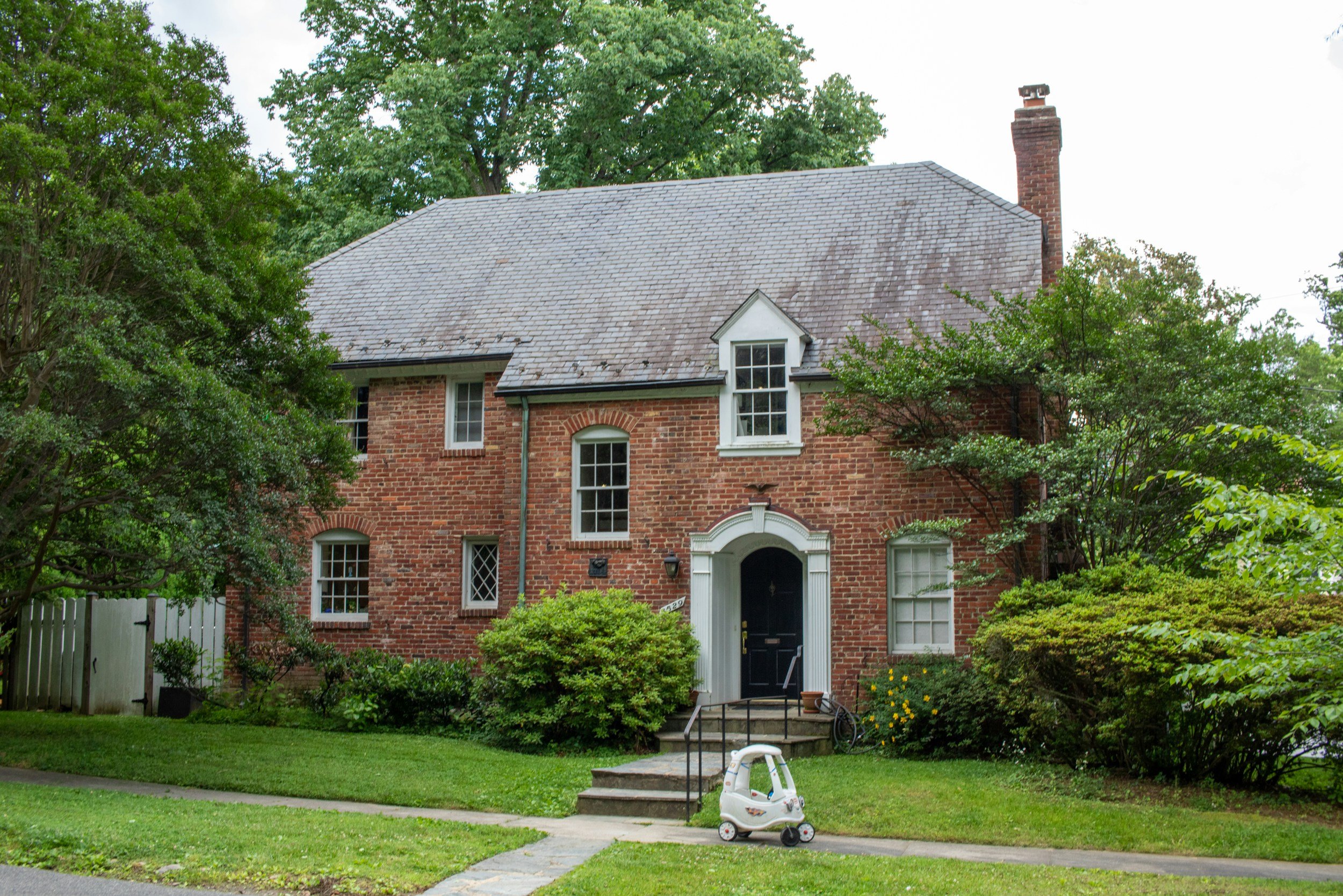 A brick house with a tall brick chimney, surrounded by green trees and bushes, with a small front yard and a child's toy car on the driveway.