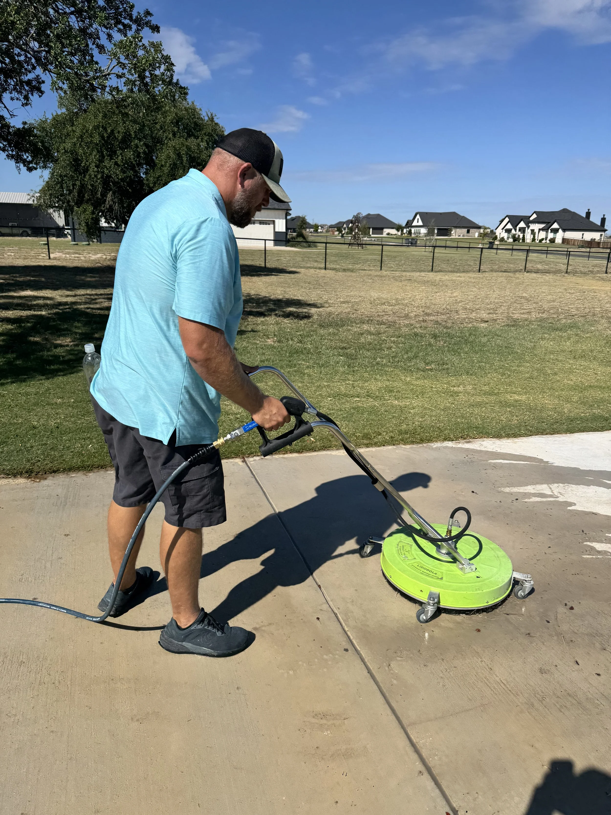 Man in blue shirt and black shorts cleaning a concrete sidewalk with a floor scrubber machine outdoors on a sunny day.