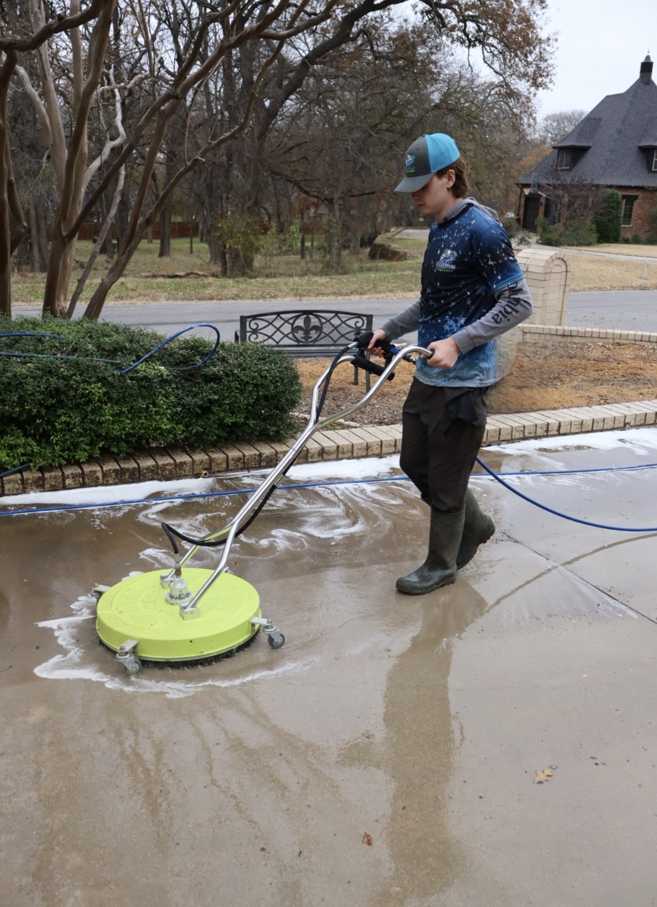 A person wearing a blue and gray cap, a blue shirt, gray long sleeves, and black rubber boots is pressure washing a concrete driveway. The driveway has soap and water on it from the cleaning. There are trees, a street, a bench, and a house with a gray roof and brick walls in the background.