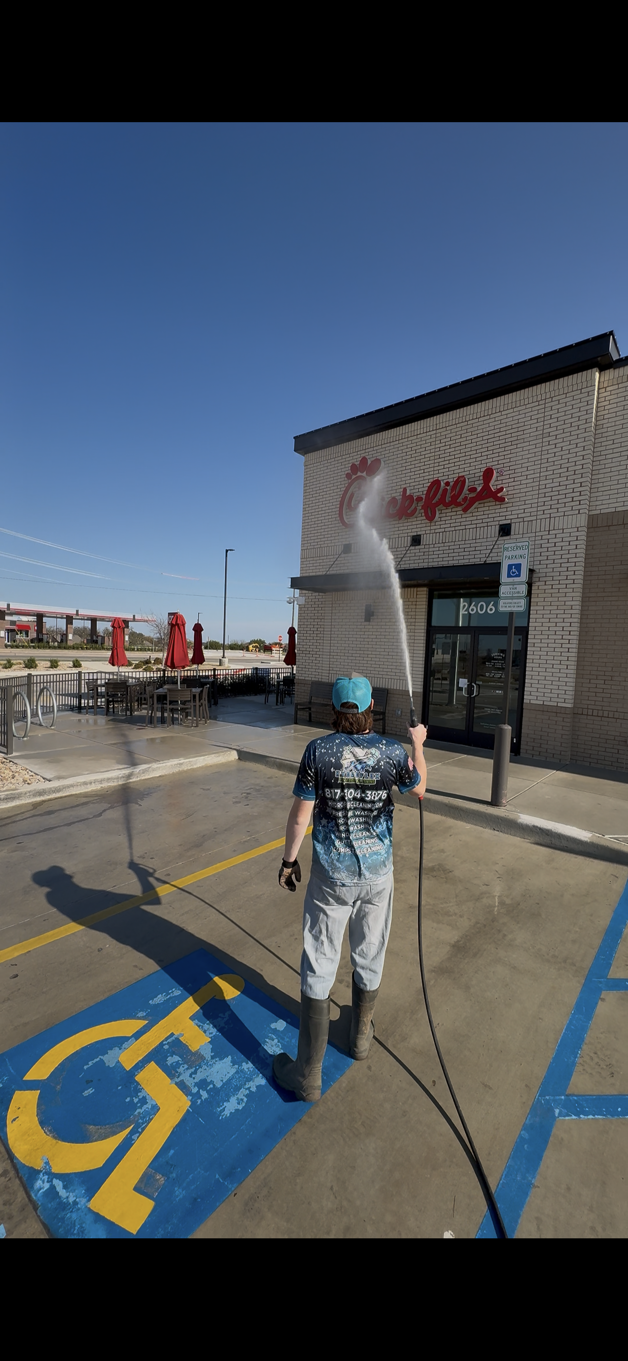 A person wearing a blue cap and a dark shirt, which displays text about washing and cleaning, is holding a high-pressure hose and spraying water outside a Chick-fil-A restaurant. The establishment has outdoor seating with red umbrellas and a reserved