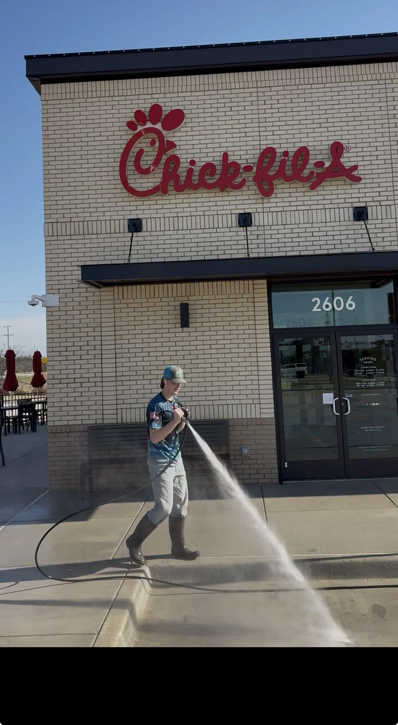 A person wearing a cap and rain boots is pressure washing the sidewalk outside a Chick-fil-A restaurant during the daytime.