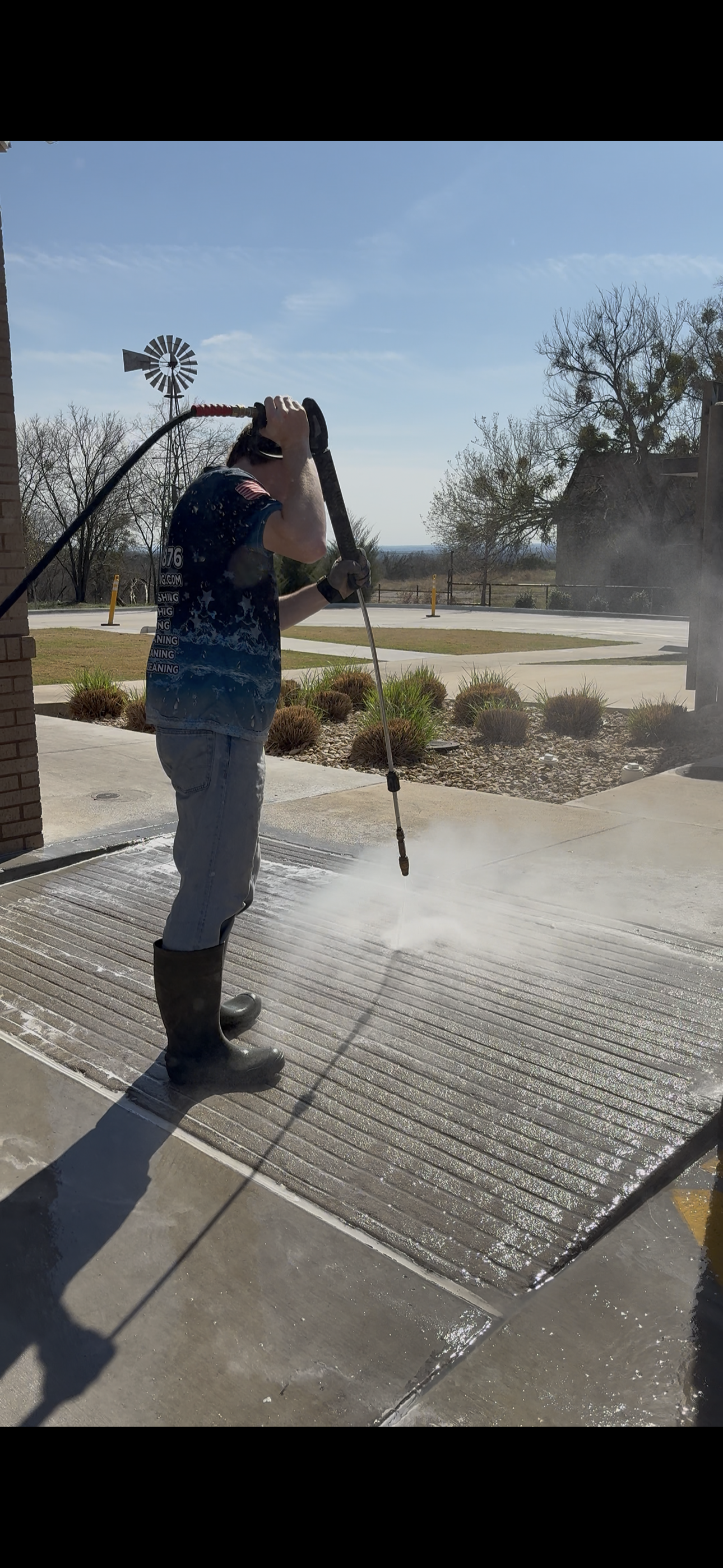A person wearing rubber boots pressure washing a concrete porch outside on a sunny day.