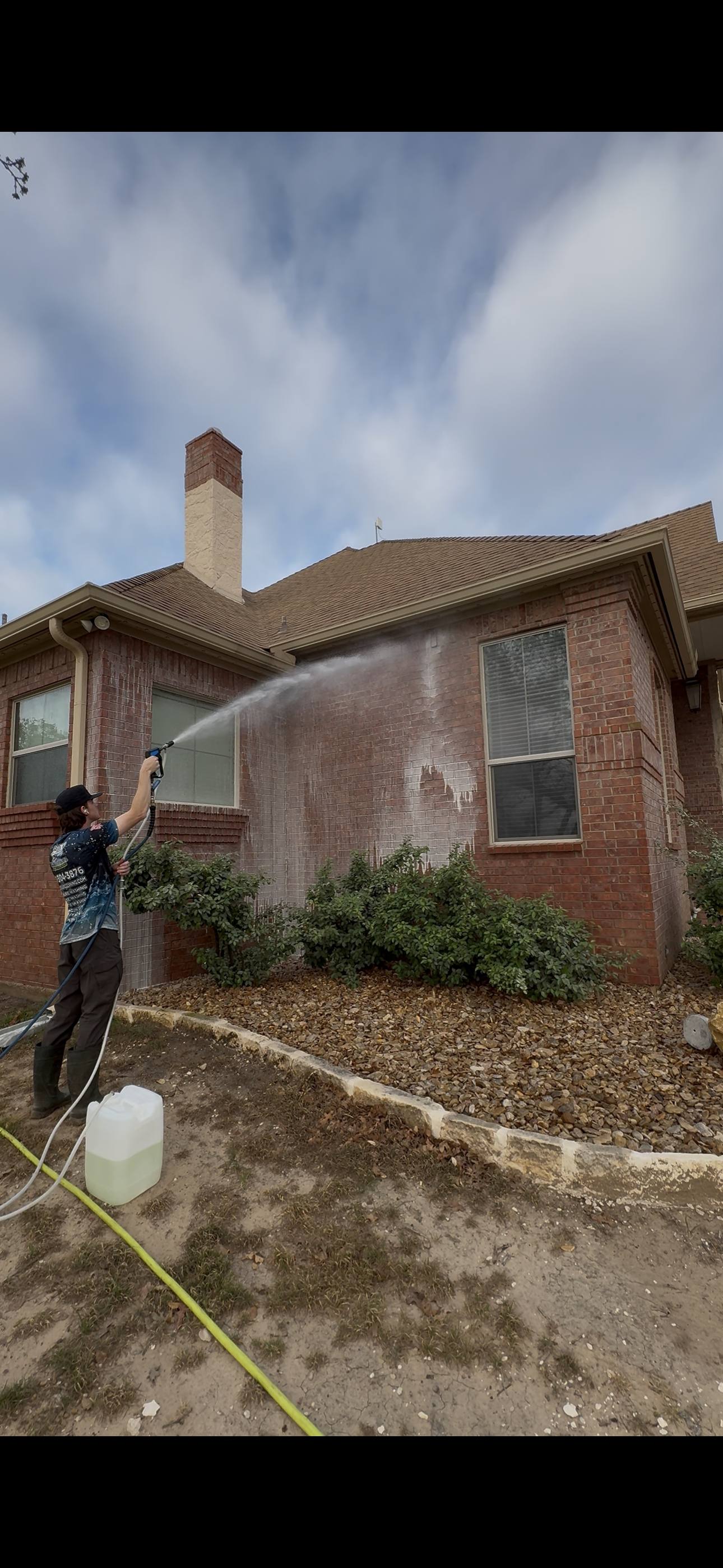A person is pressure washing the brick exterior of a house with a garden and bushes in front.