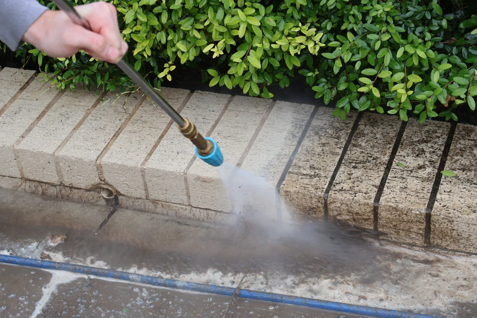 Person using a pressure washer to clean a brick curb near green shrubs.