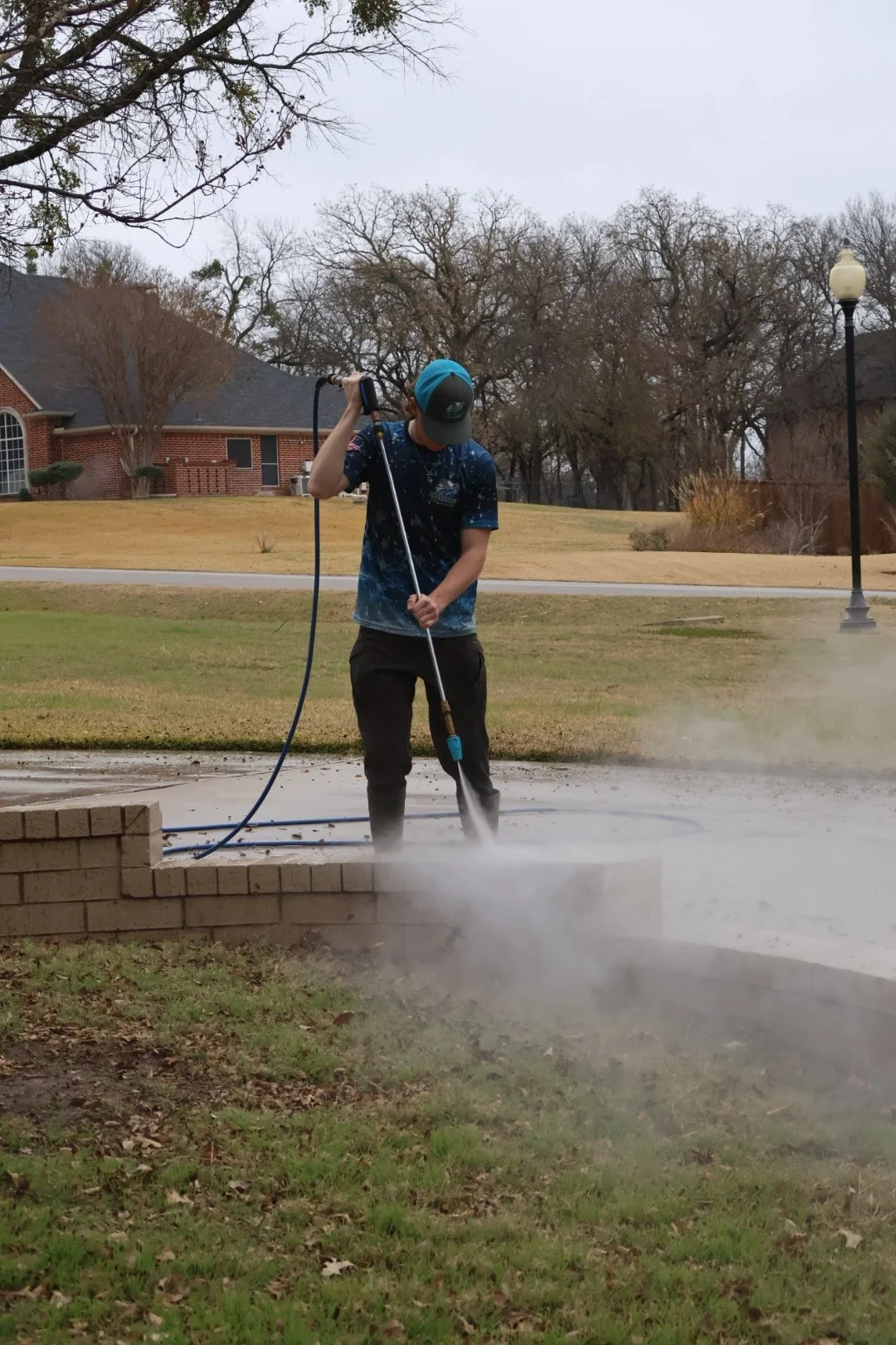 A person pressure washing a concrete sidewalk outdoors, wearing a baseball cap and dark pants, with a house, trees, and a street lamp in the background on a cloudy day.