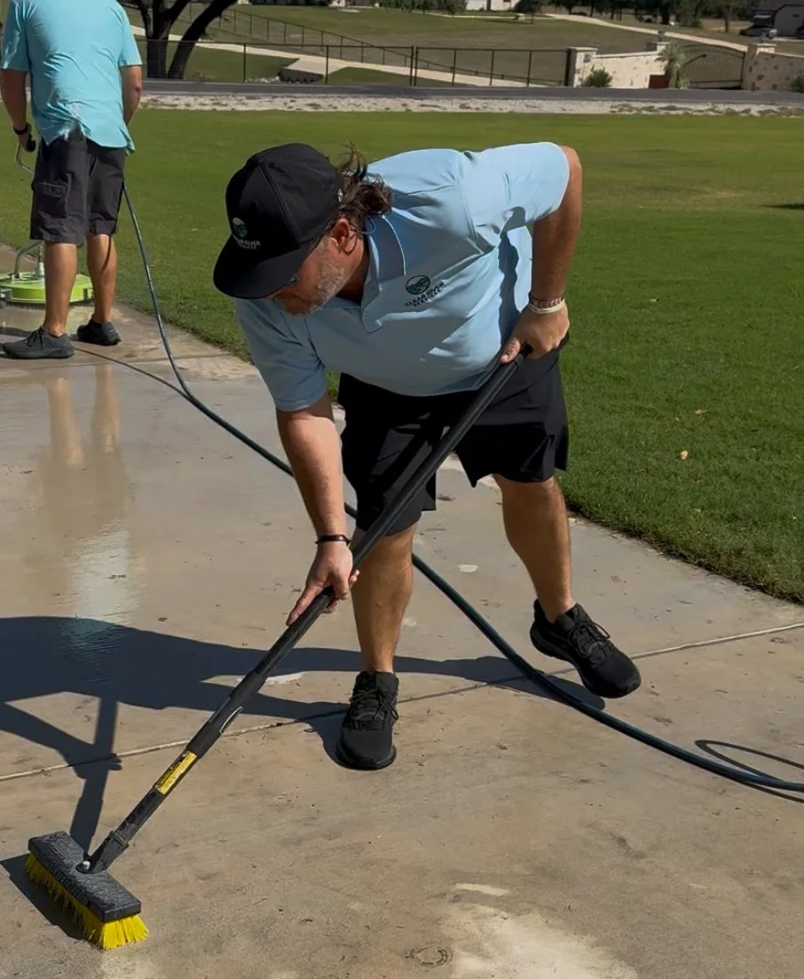 A man with long hair, wearing a black cap, light blue polo, and black shorts, is bending down and sweeping a concrete surface with a push broom. Another person in the background is standing on the edge of the concrete, holding a hose. The setting app