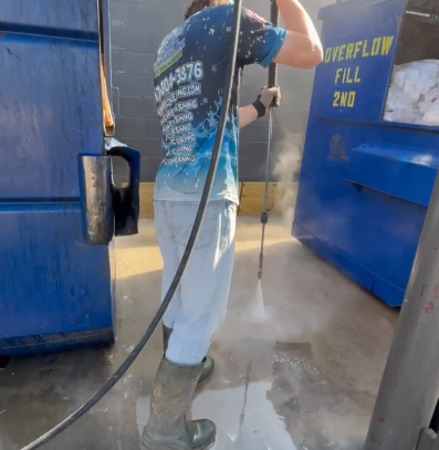Person using a pressure washer to clean a concrete surface near large blue dumpsters.