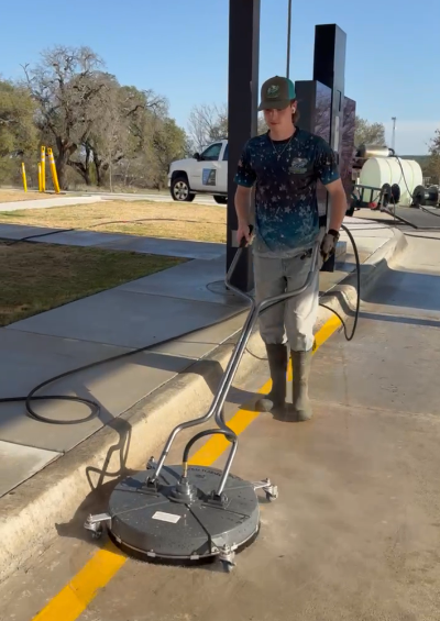 Worker uses a ride-on floor scrubber machine to clean a concrete parking lot. He wears a cap, gloves, gray pants, and tall black boots.