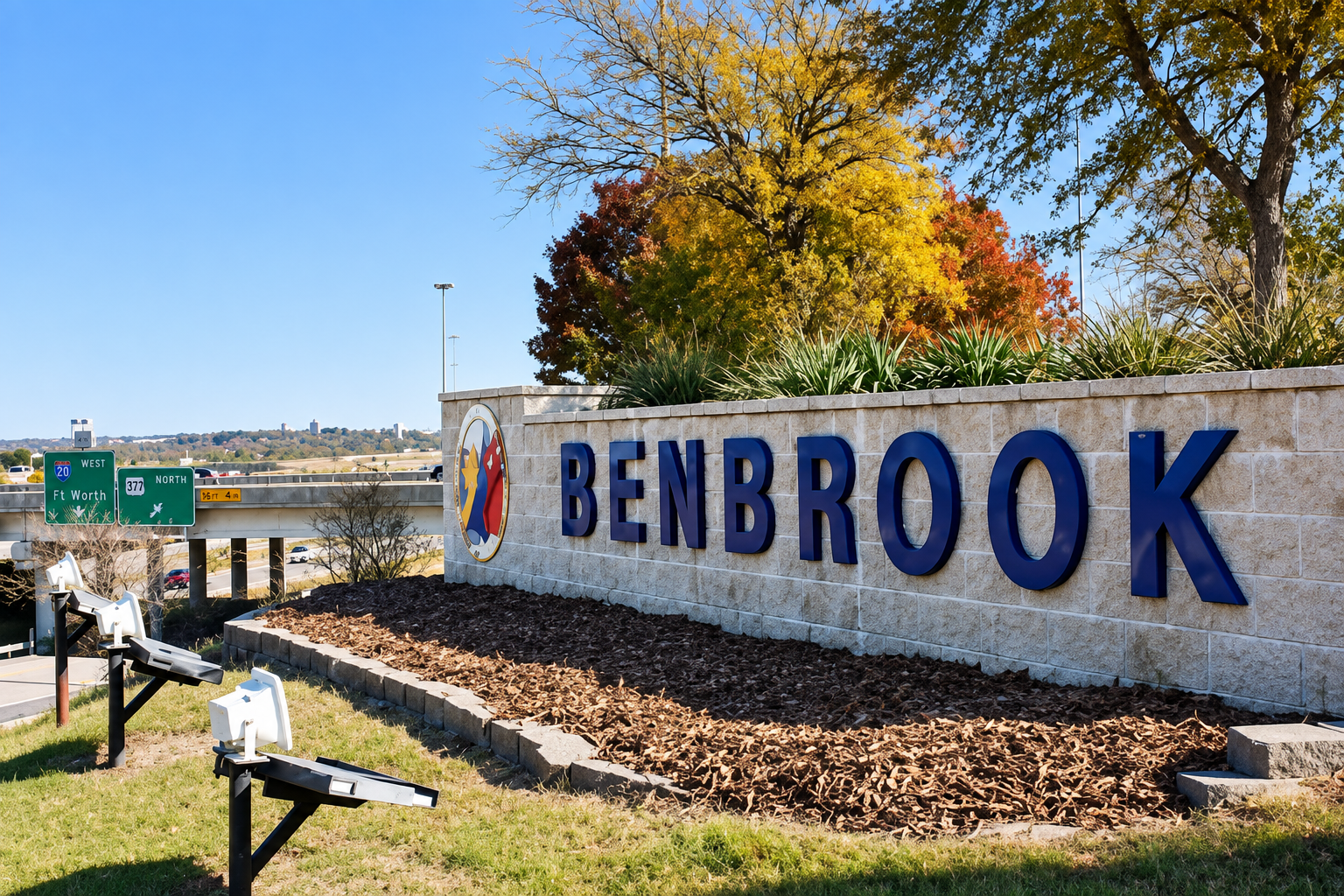 Entrance sign for Benbrook, Texas, with large blue letters on a stone wall, surrounded by autumn-colored trees and a landscaped area with mulch.