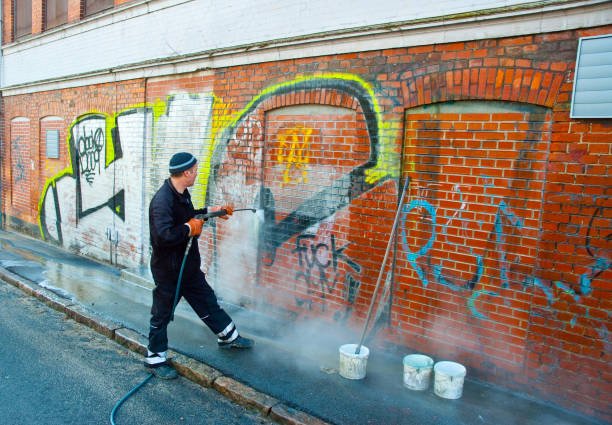 A man in dark clothing and a beanie cleaning graffiti off a brick wall with a pressure washer, next to buckets and spray foam. The wall has multiple graffiti tags in various colors.