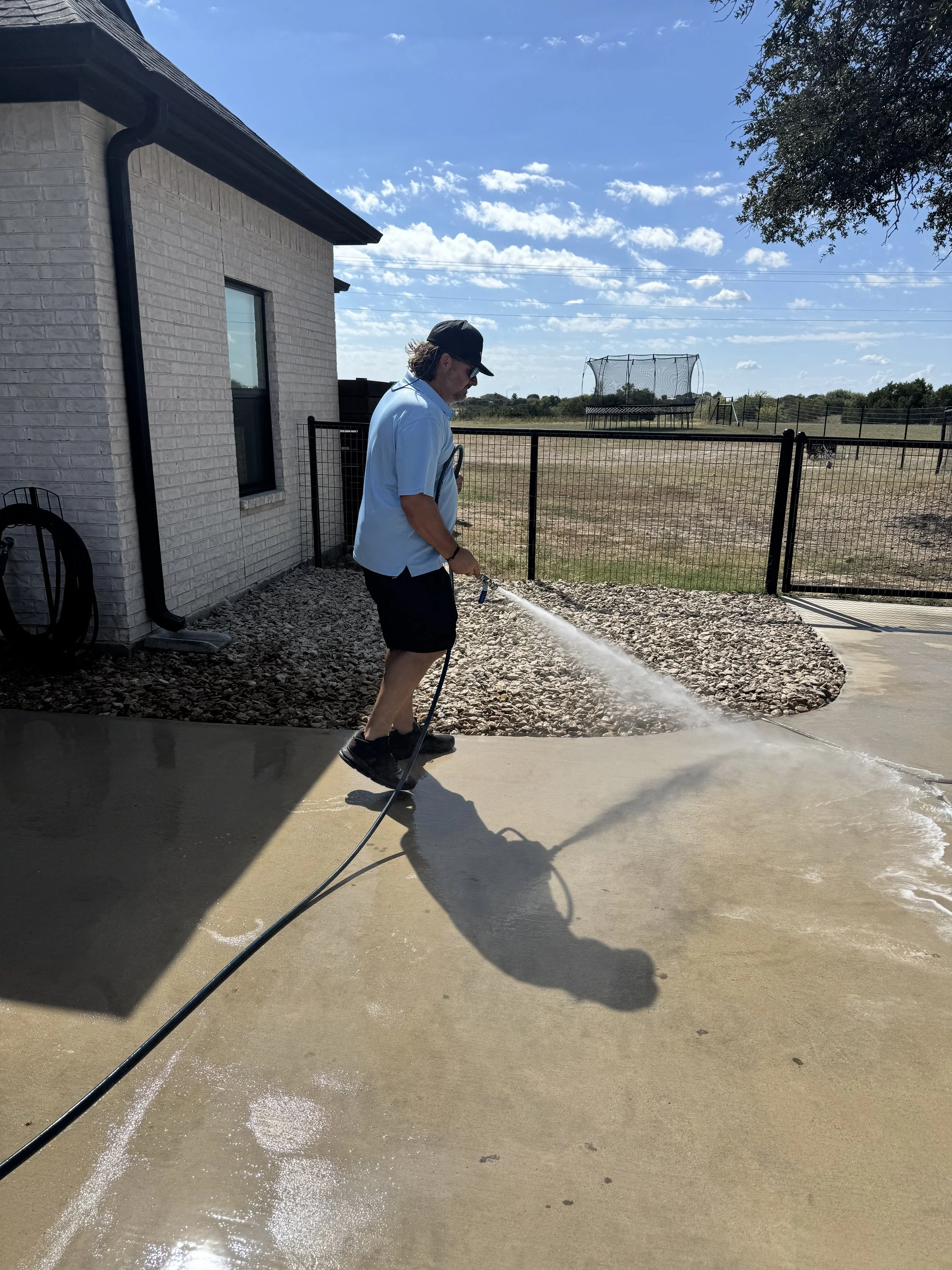 A man in a blue shirt, black shorts, a black cap, and sunglasses is watering a concrete patio with a garden hose on a sunny day. The background shows a fenced backyard with a trampoline, a grassy field, and a blue sky with some clouds.