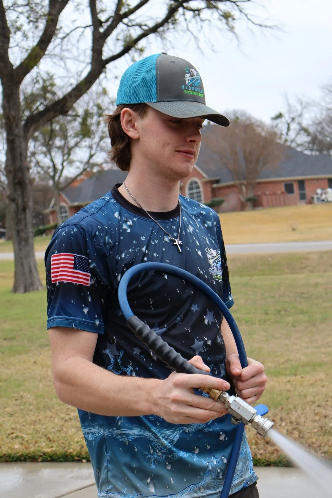 A young man wearing a blue and black cap, a blue patterned shirt with an American flag patch, and a cross necklace, is holding a garden hose sprayer with water spraying from it in a residential neighborhood yard with grass, trees, and houses in the background.