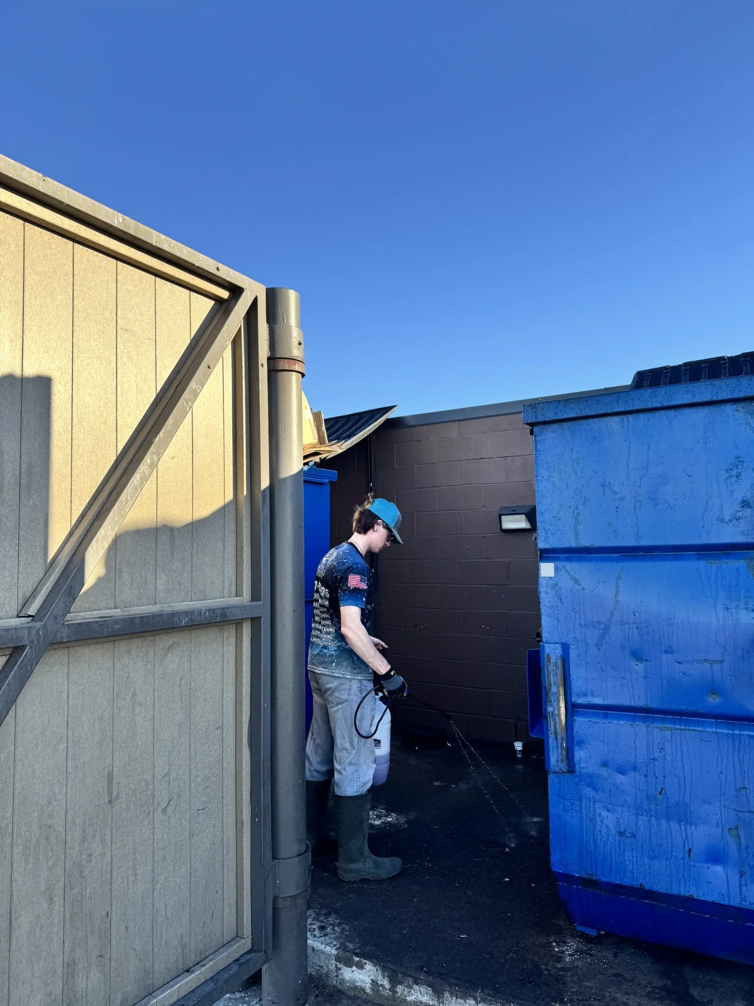 A person wearing gloves and boots, standing near a large blue dumpster, using a pressure washer to clean the ground next to a brown wall and a beige wall under a clear blue sky.