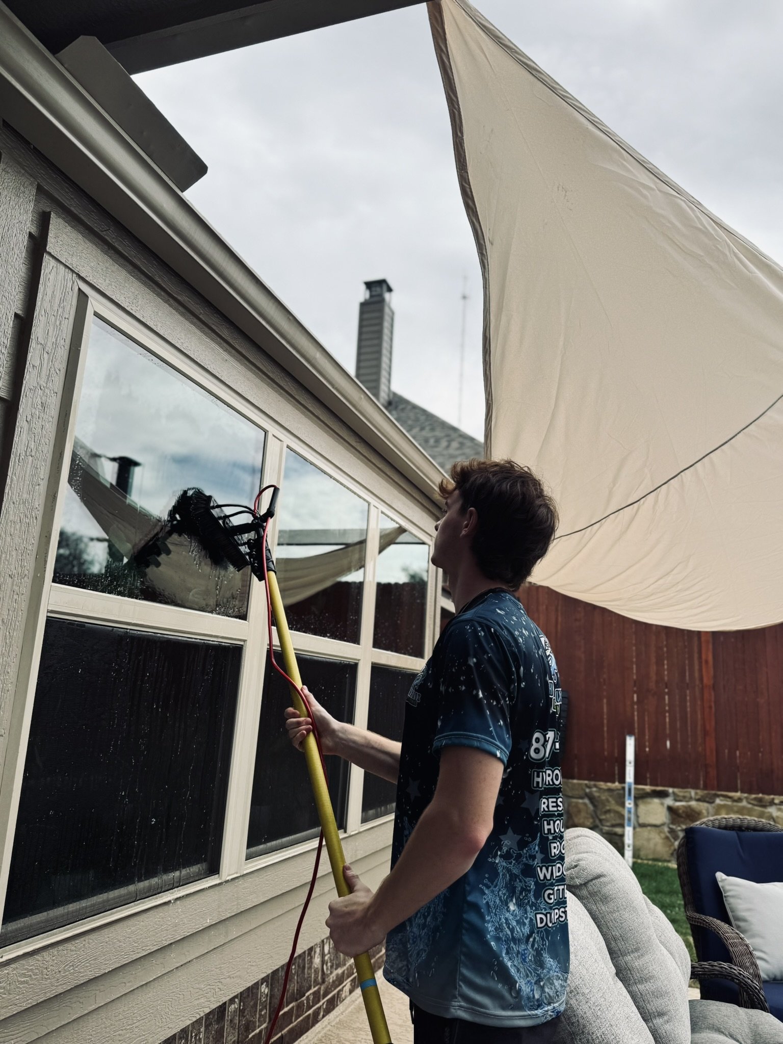 A person cleaning a window with a long-handled squeegee outside a house on a cloudy day.