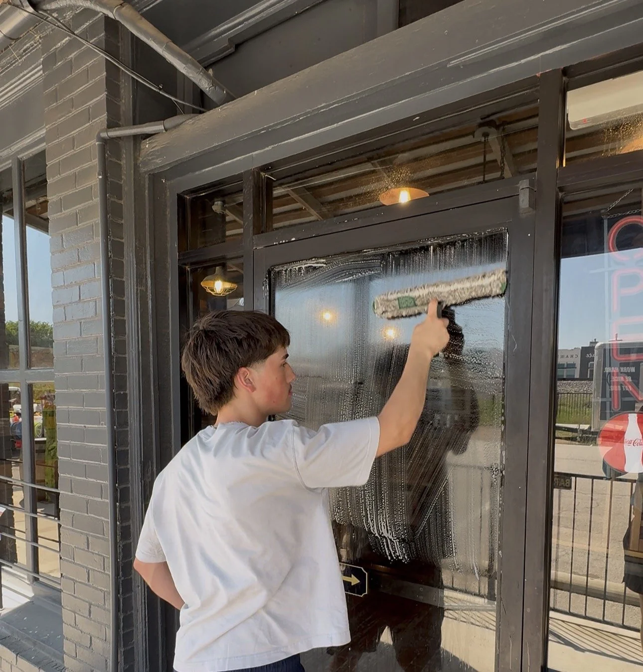 A young boy cleaning a glass door with a squeegee, outside a building with brick and metal framing.