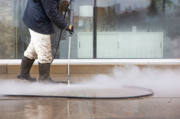 Person in black raincoat and rubber boots using steam cleaner on sidewalk outside building.