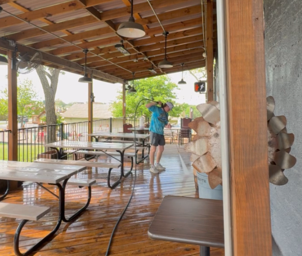 Empty outdoor patio with wooden flooring, hanging lights, and metal picnic tables, with a person in a blue shirt pouring a drink on a wooden deck under a wooden roof.