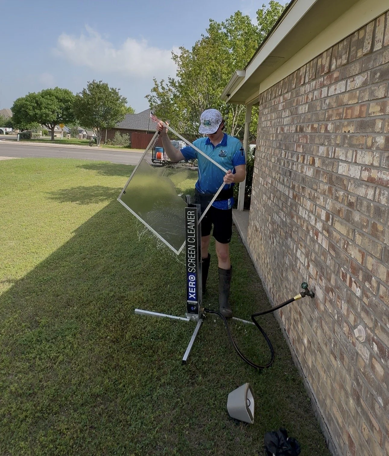A person standing outdoors next to a brick house, wearing a blue shirt, black shorts, and rain boots, filling a transparent anti-graffiti screen with water from a hose. The screen is mounted on a stand labeled "XERO SCREEN CLEANER" and appears to be 